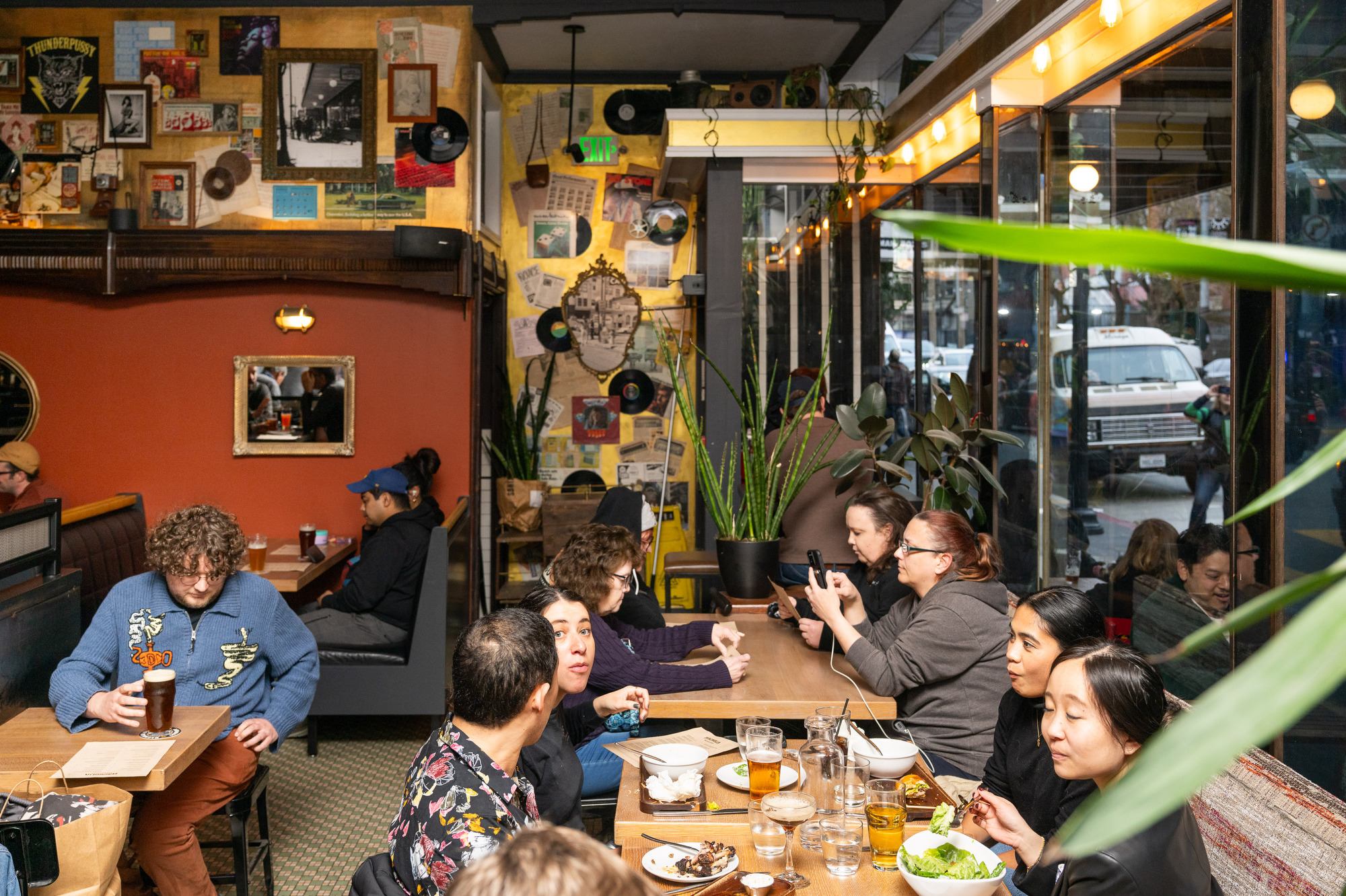 A lively cafe scene features people chatting and eating, surrounded by eclectic wall decor like photos and vinyl records. Large windows let in natural light.