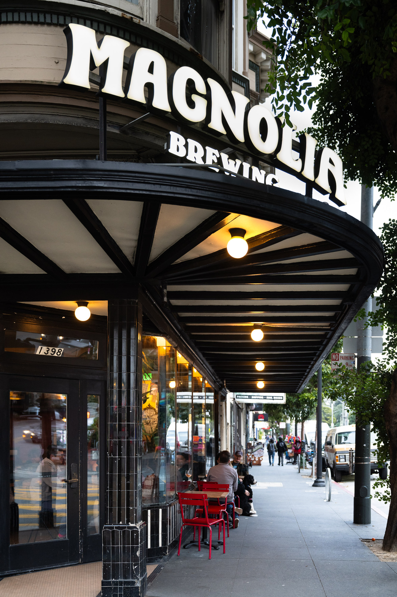 A sidewalk view of a brewery with black and white awning and a sign reading &quot;Magnolia Brewing.&quot; Red chairs are outside, and people are seated and walking.