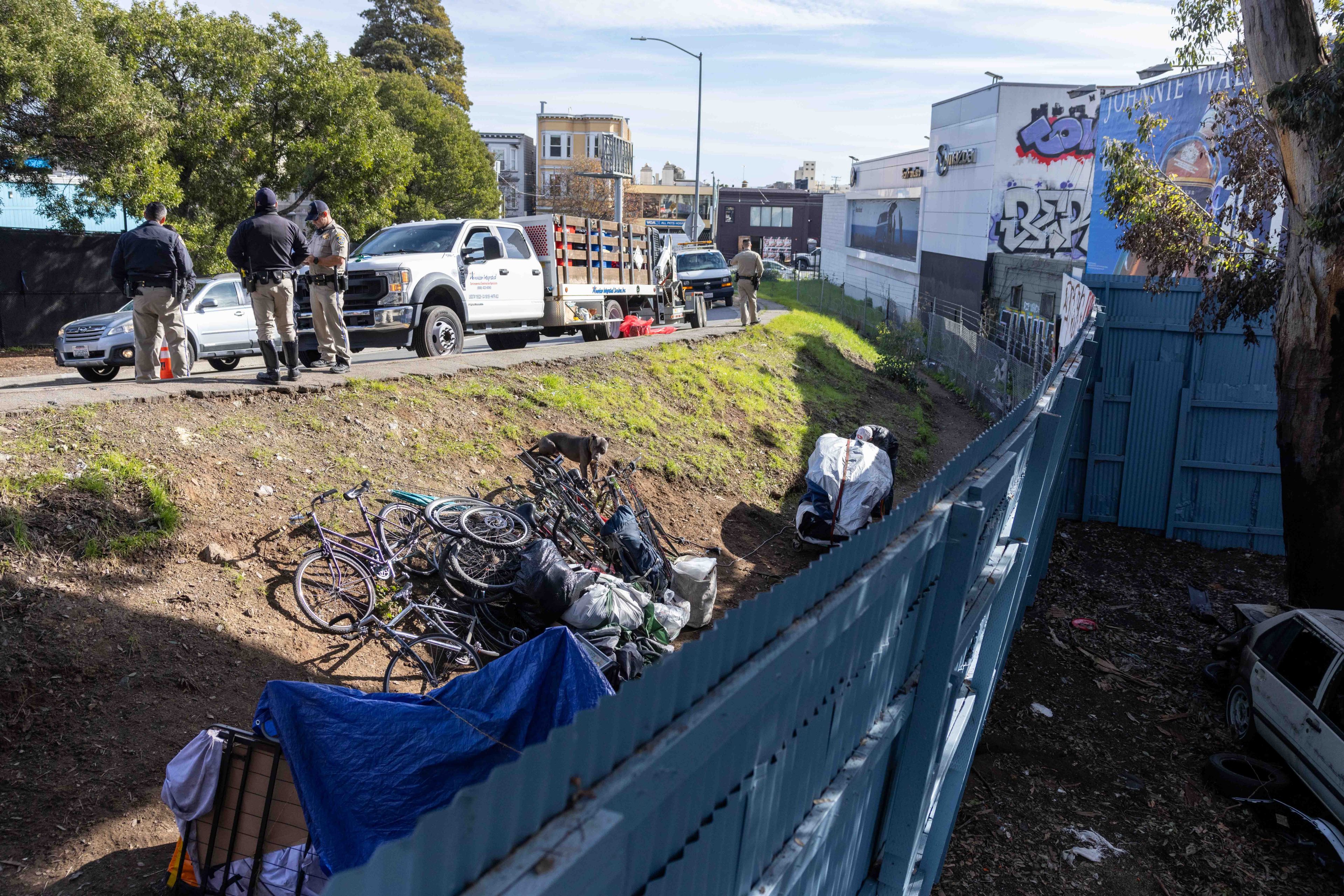 In the image, several officials stand near a pickup truck on a roadside. Below them, a makeshift camp with bikes and tarps is visible on a grassy slope. A dog is nearby.