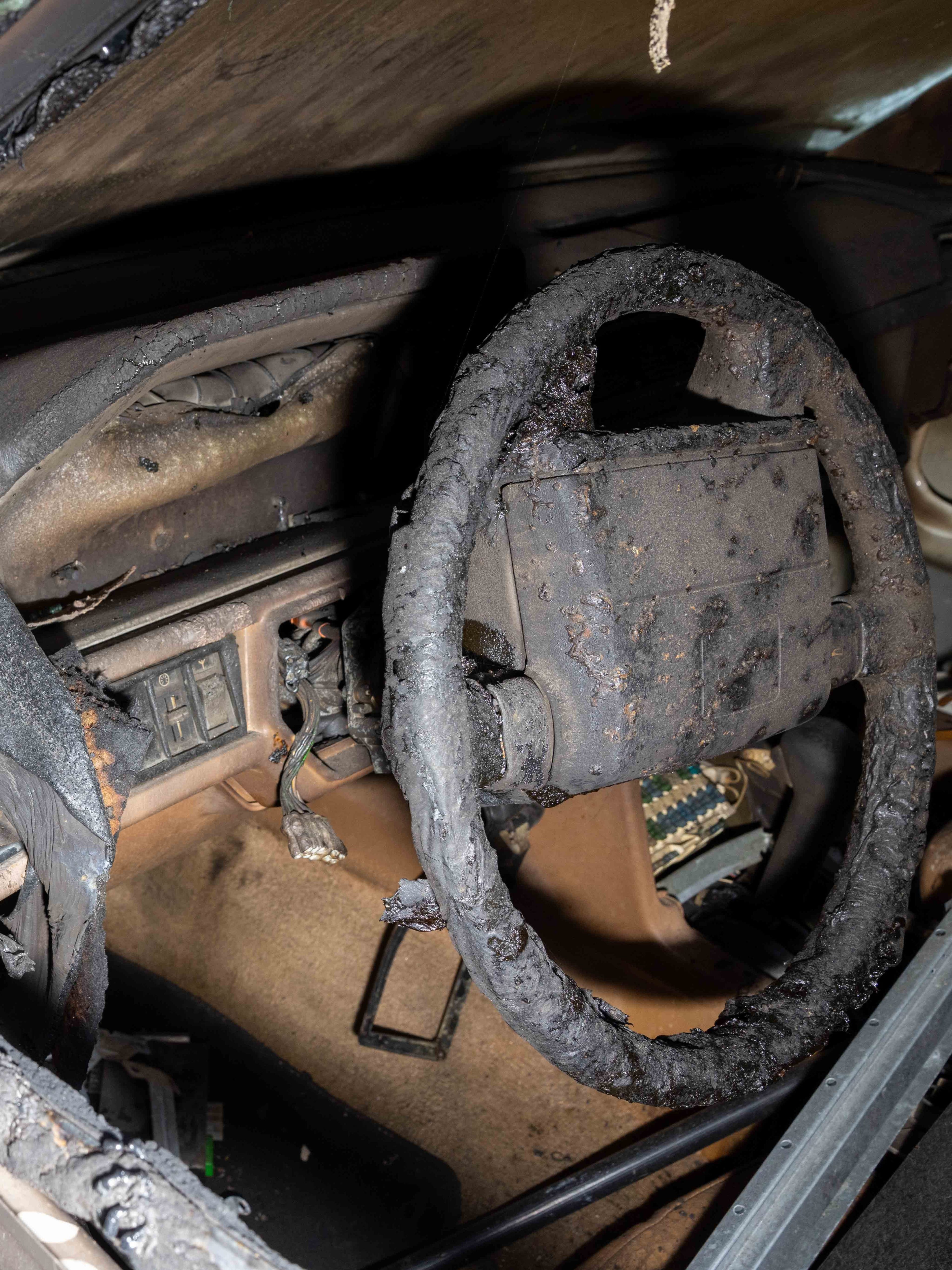 The image shows a heavily burned car interior with the steering wheel and dashboard covered in soot and charred remnants. Wires are exposed, and the floor is scorched.