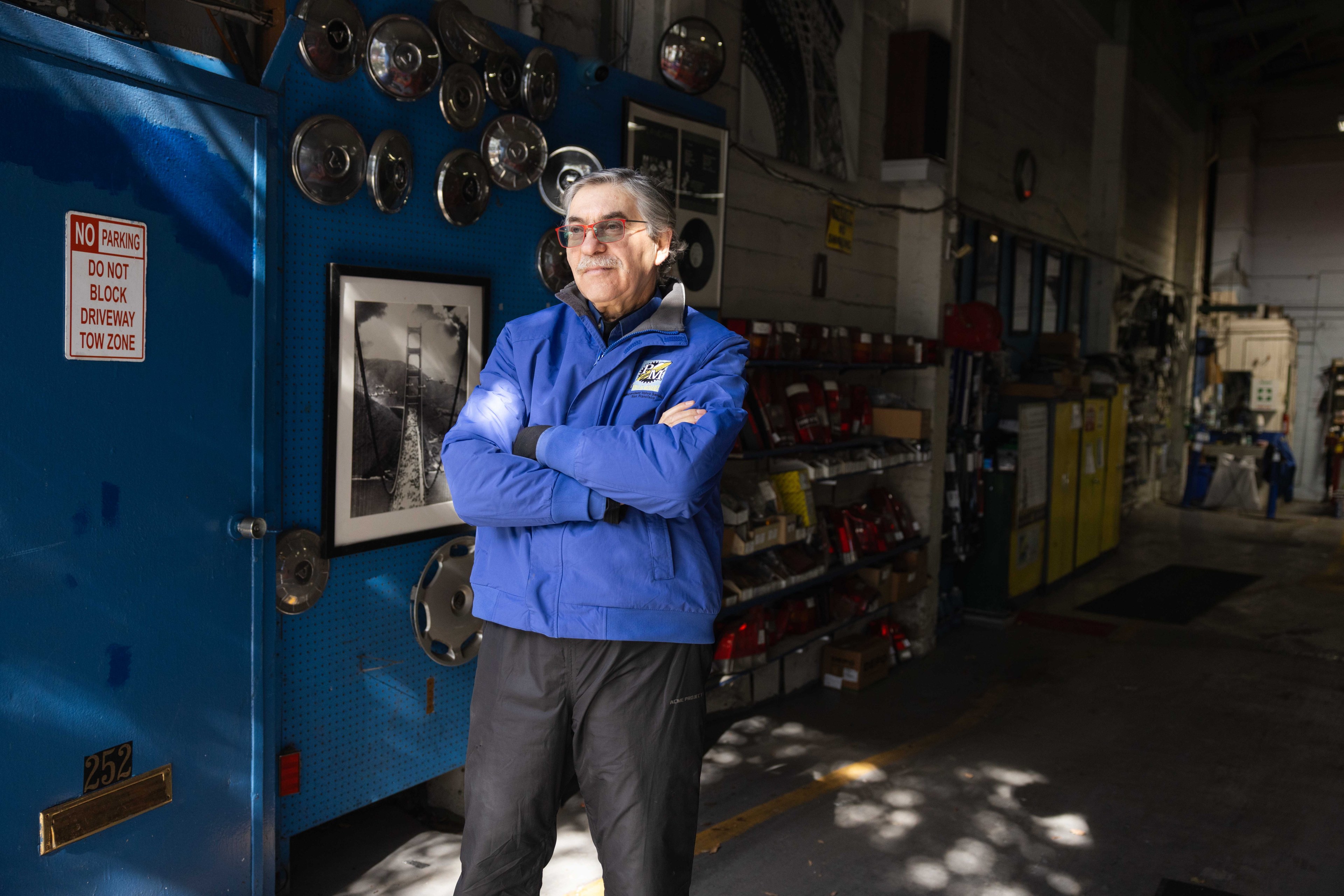 A person in a blue jacket stands with crossed arms in a workshop. Behind them, hubcaps are displayed on a wall, along with a black-and-white photo. Shelves are to the right.