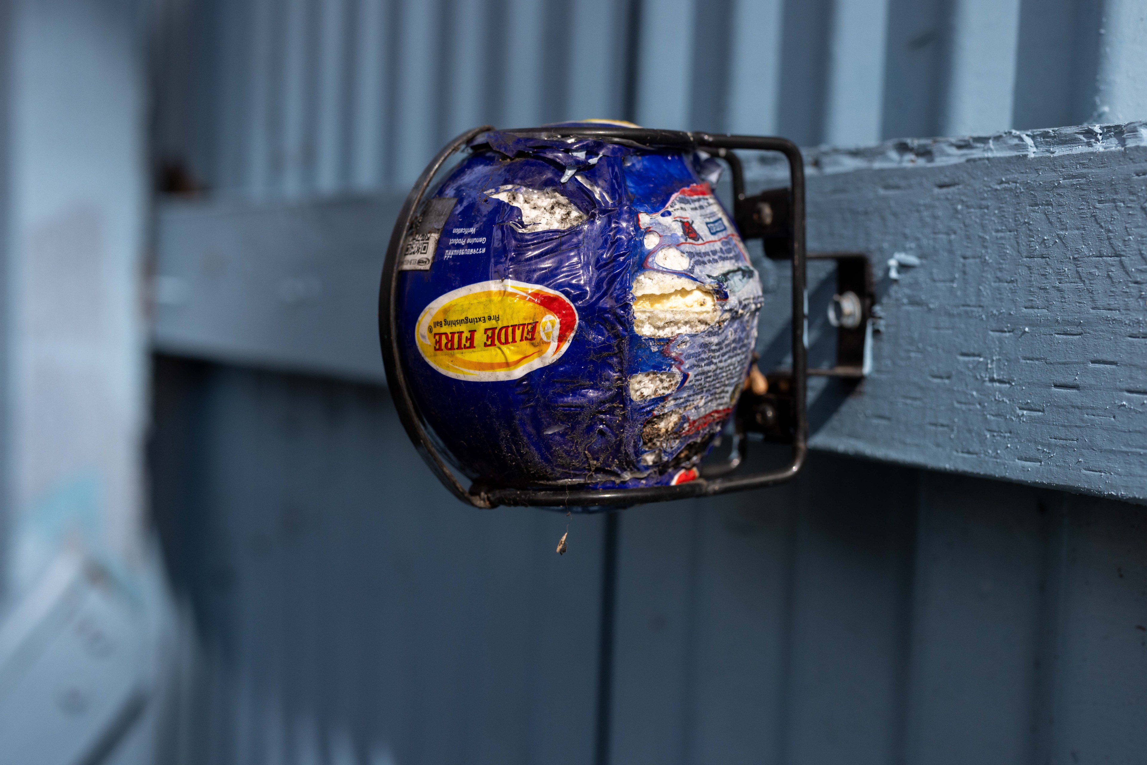 A damaged blue fire extinguisher ball is mounted on a blue metal wall. It has visible dents and exposed inner material, with a prominent yellow label.