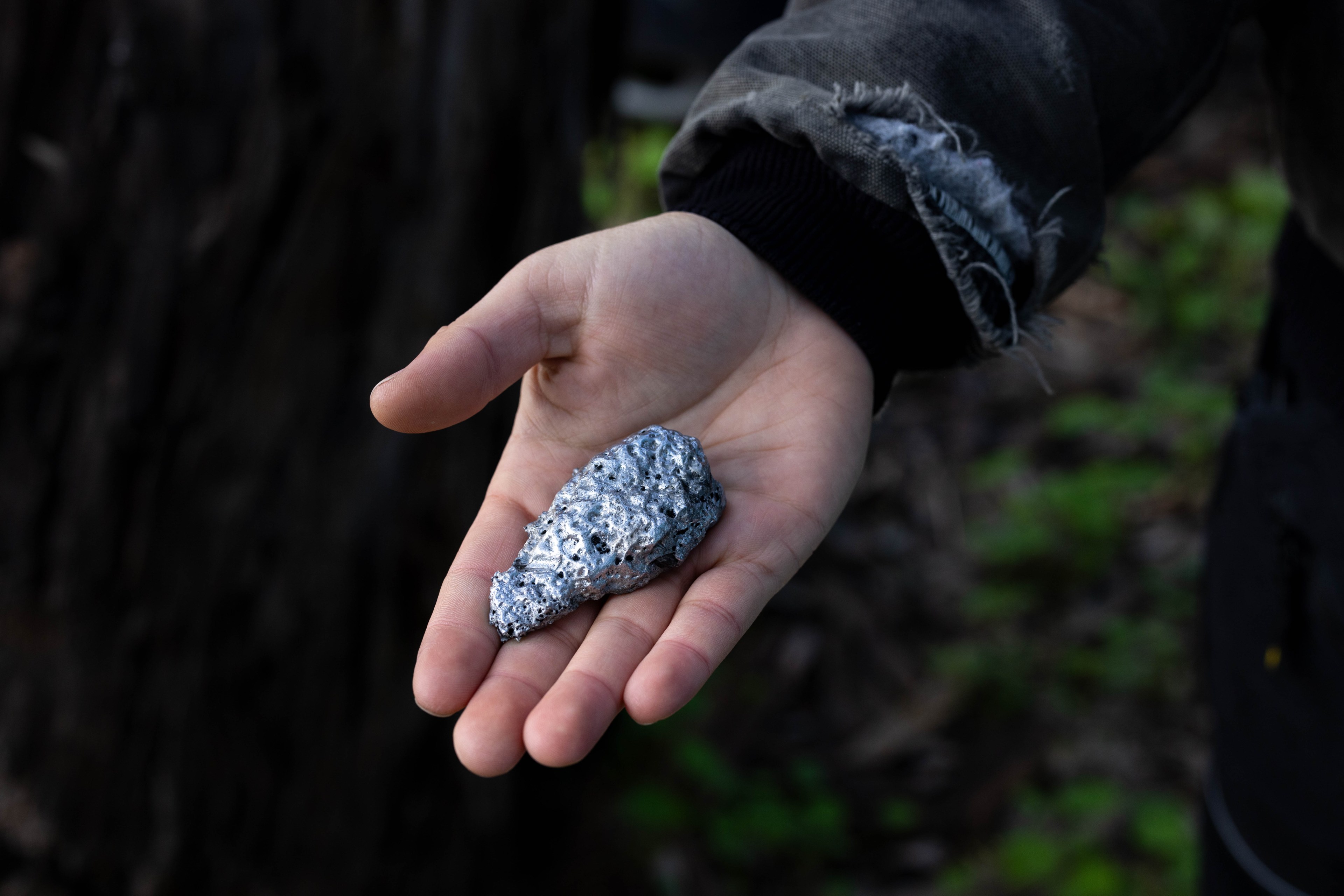 A person holds a textured, irregular piece of metallic-looking rock in their open palm, with a frayed sleeve visible, against a blurred outdoor background.