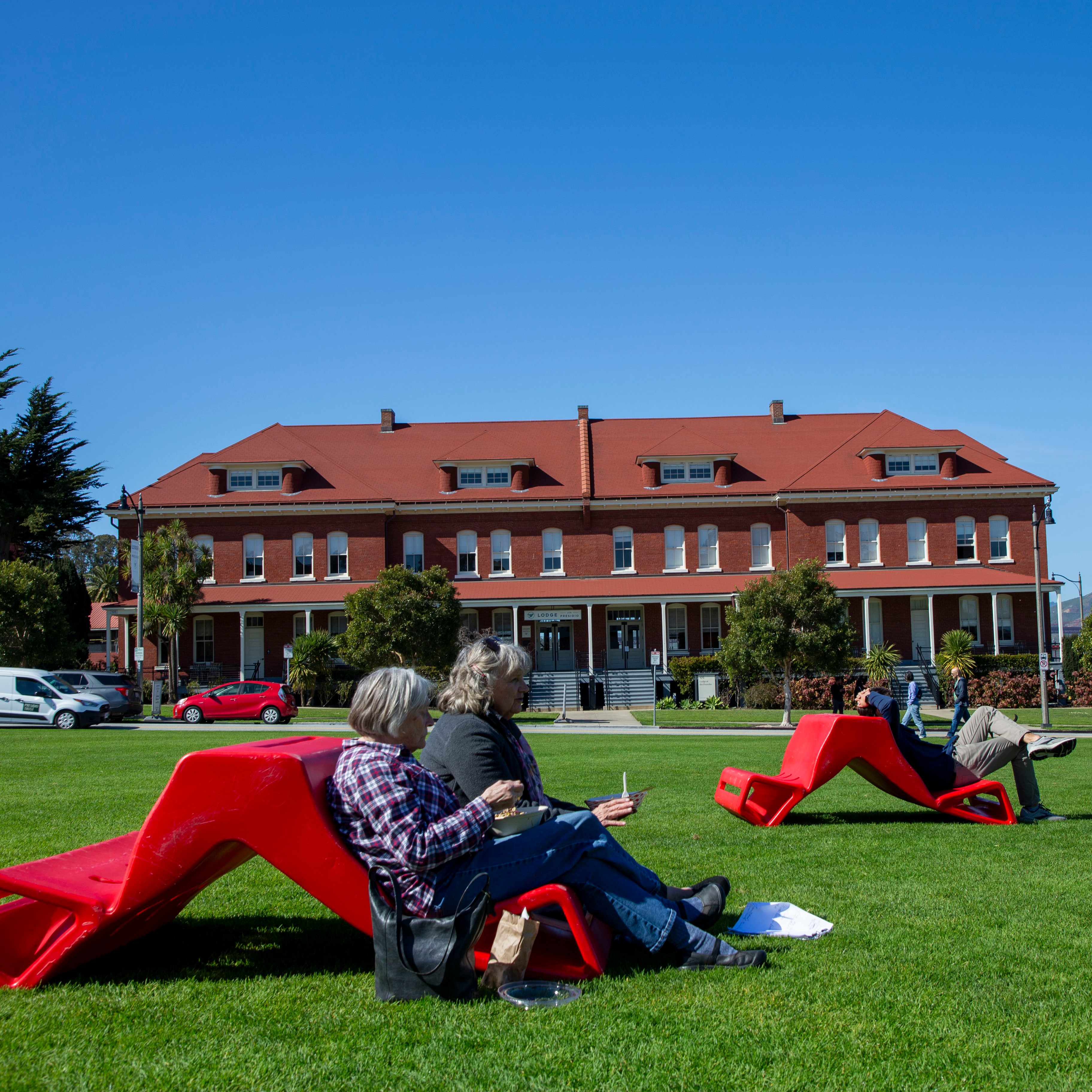 People are sitting on red lounge chairs on a grassy lawn in front of a large, red-roofed brick building under a clear blue sky.