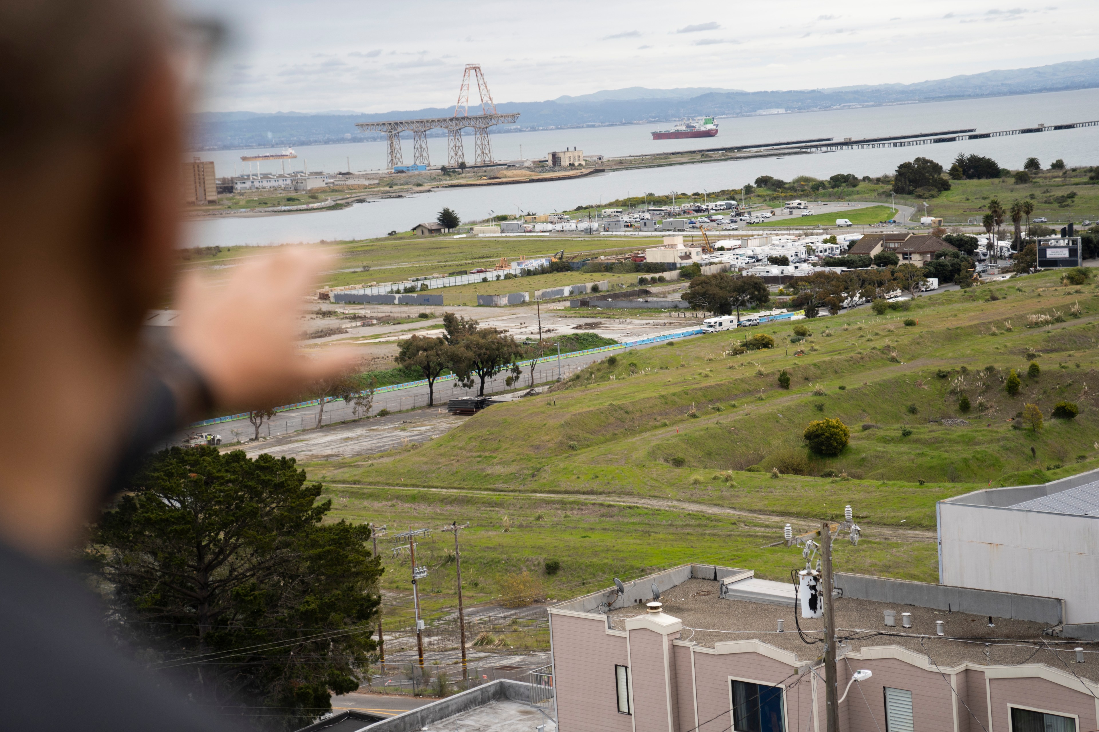 A person points towards a landscape featuring grassy hills, a waterfront, industrial structures, and a distant ship under a cloudy sky.