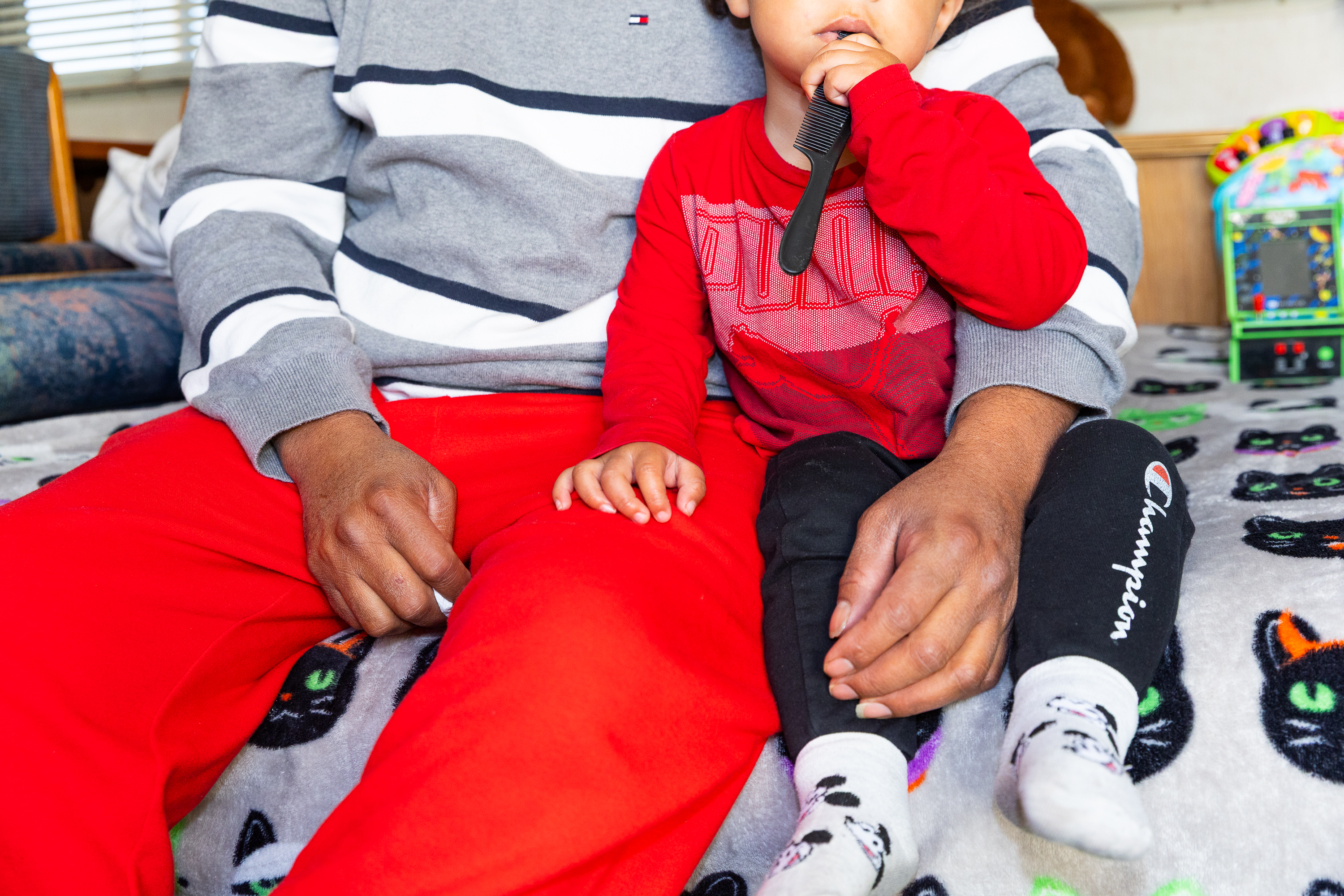 A person in a gray striped sweater and red pants sits beside a child in a red shirt and black pants who holds a comb. They're on a blanket with cat designs.