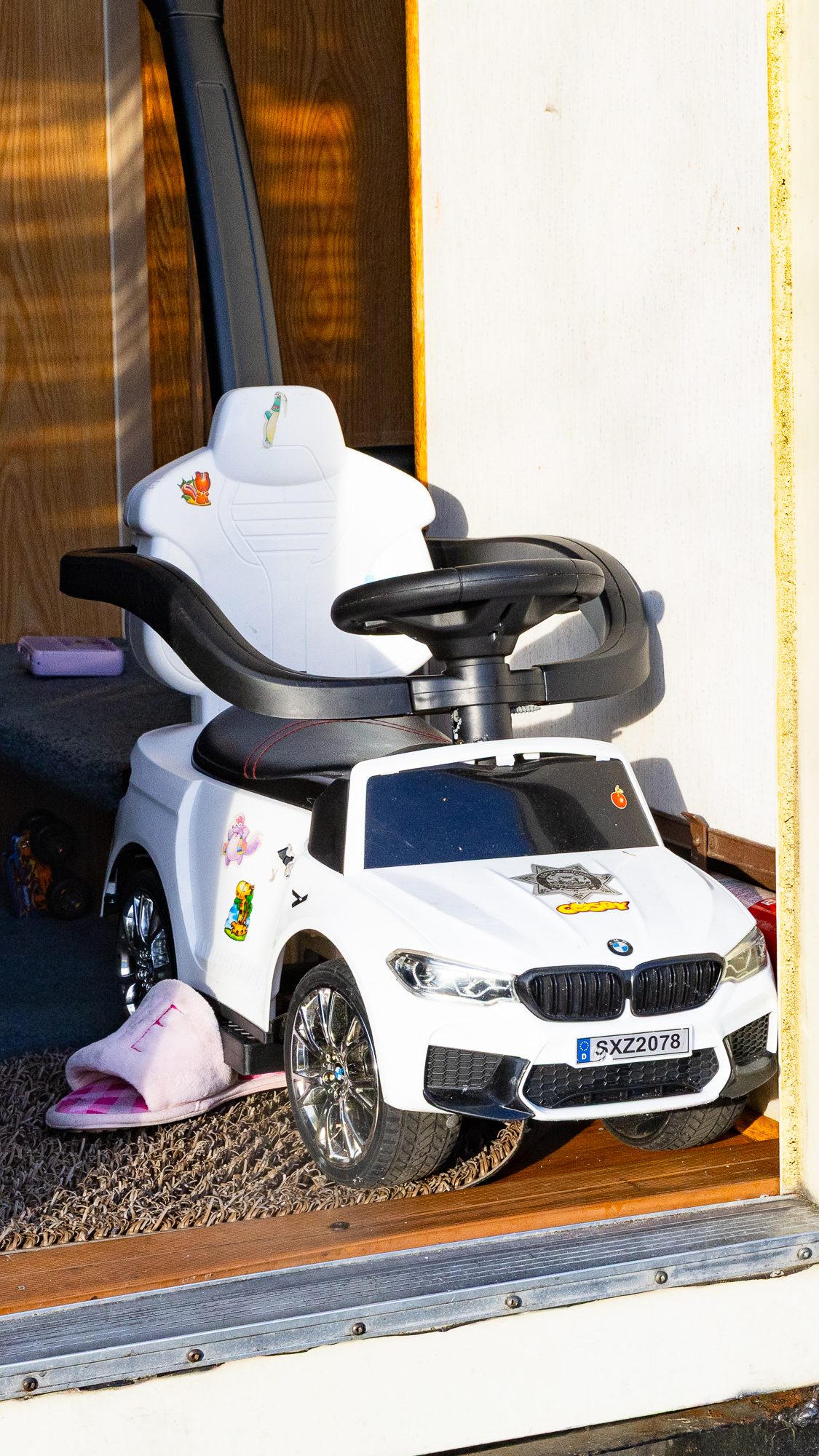 A white toy car resembling a luxury vehicle is parked indoors. It’s adorned with fun stickers, and a pink slipper rests nearby on a textured mat.