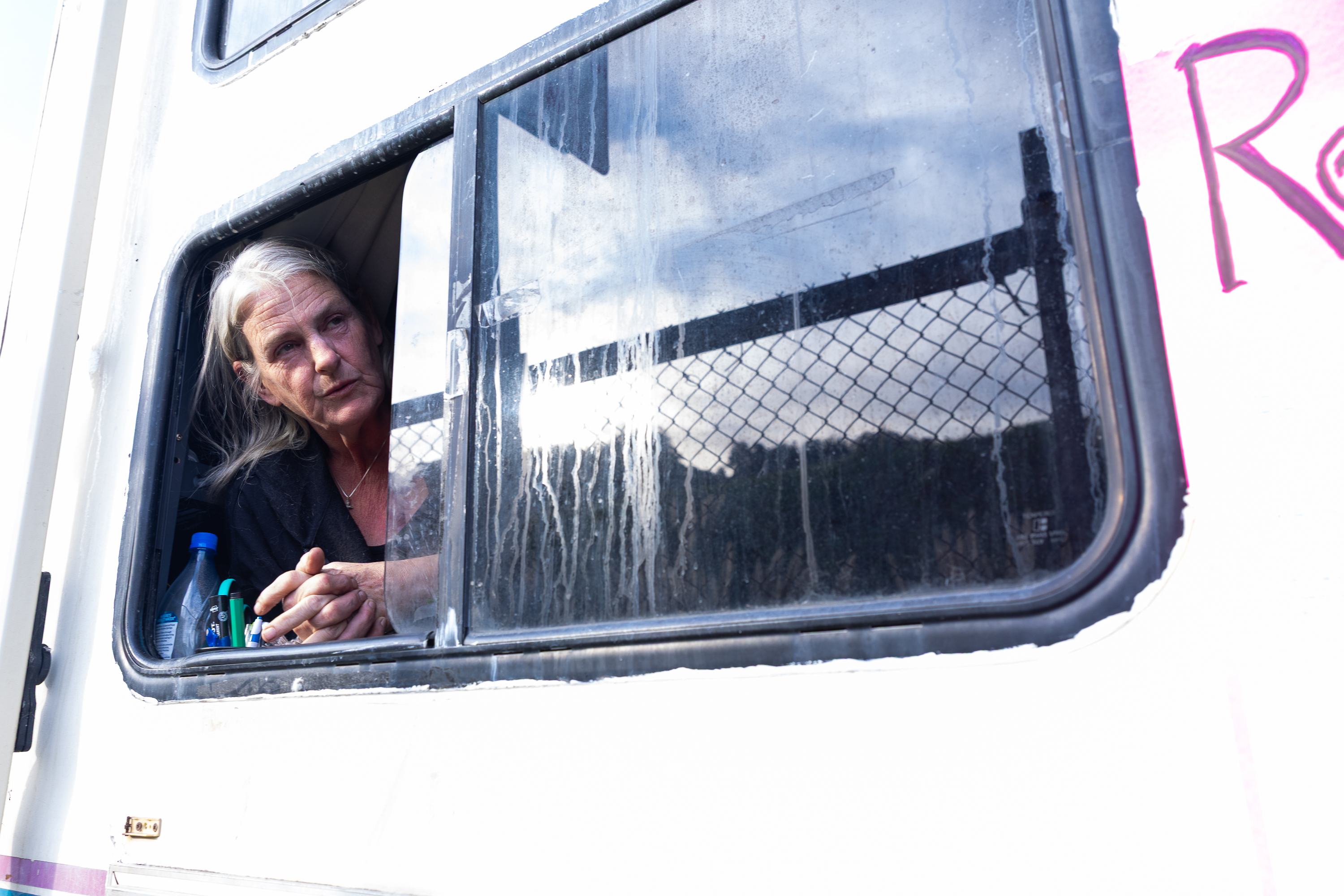 A person with long hair leans on a dirty window from inside a vehicle, holding something in their hands. A chain-link fence and cloudy sky appear outside.