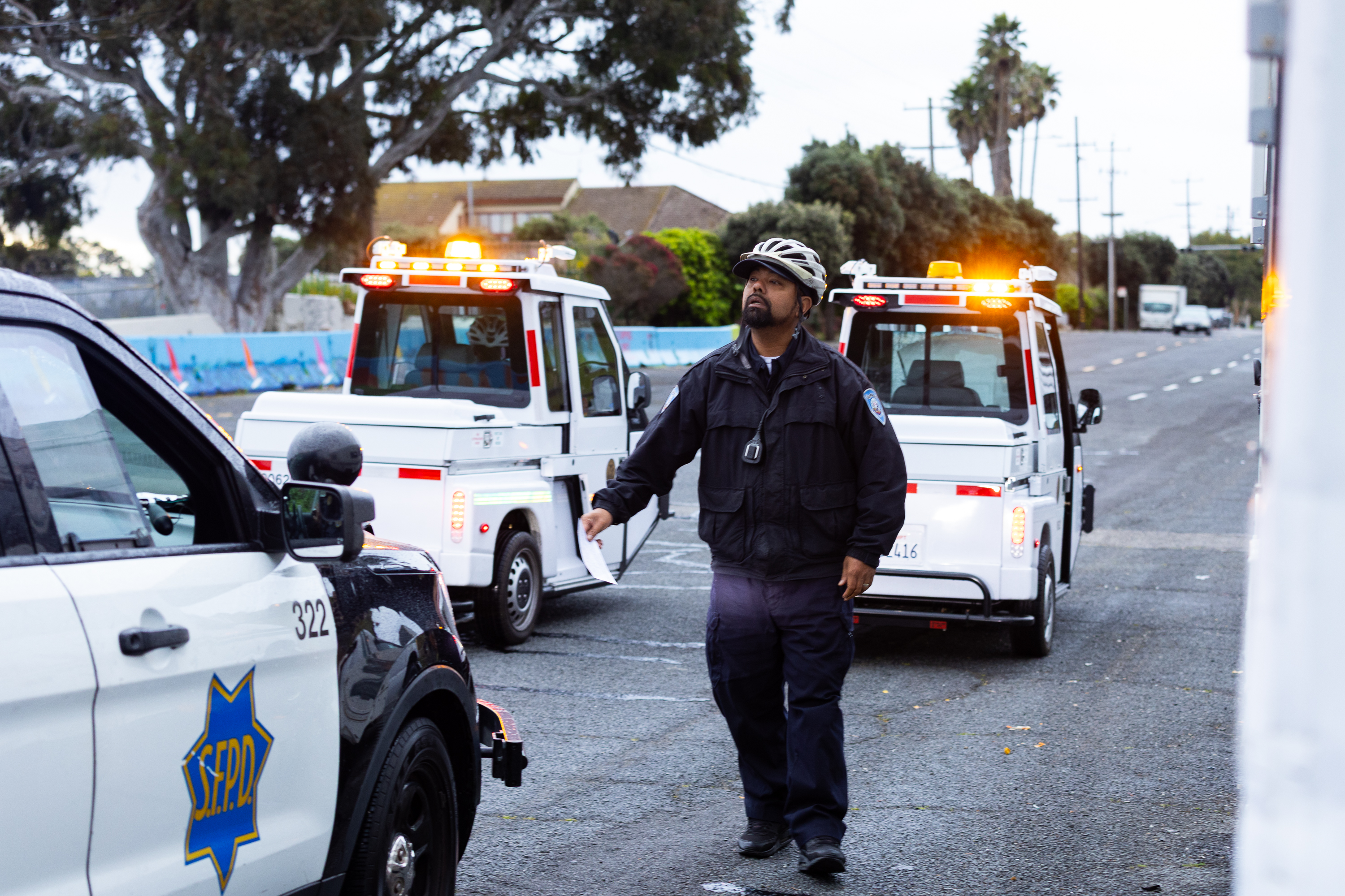 A police officer wearing a helmet stands near a patrol car with &quot;SFPD&quot; on the door. Two small vehicles with orange lights are in the background on a street.