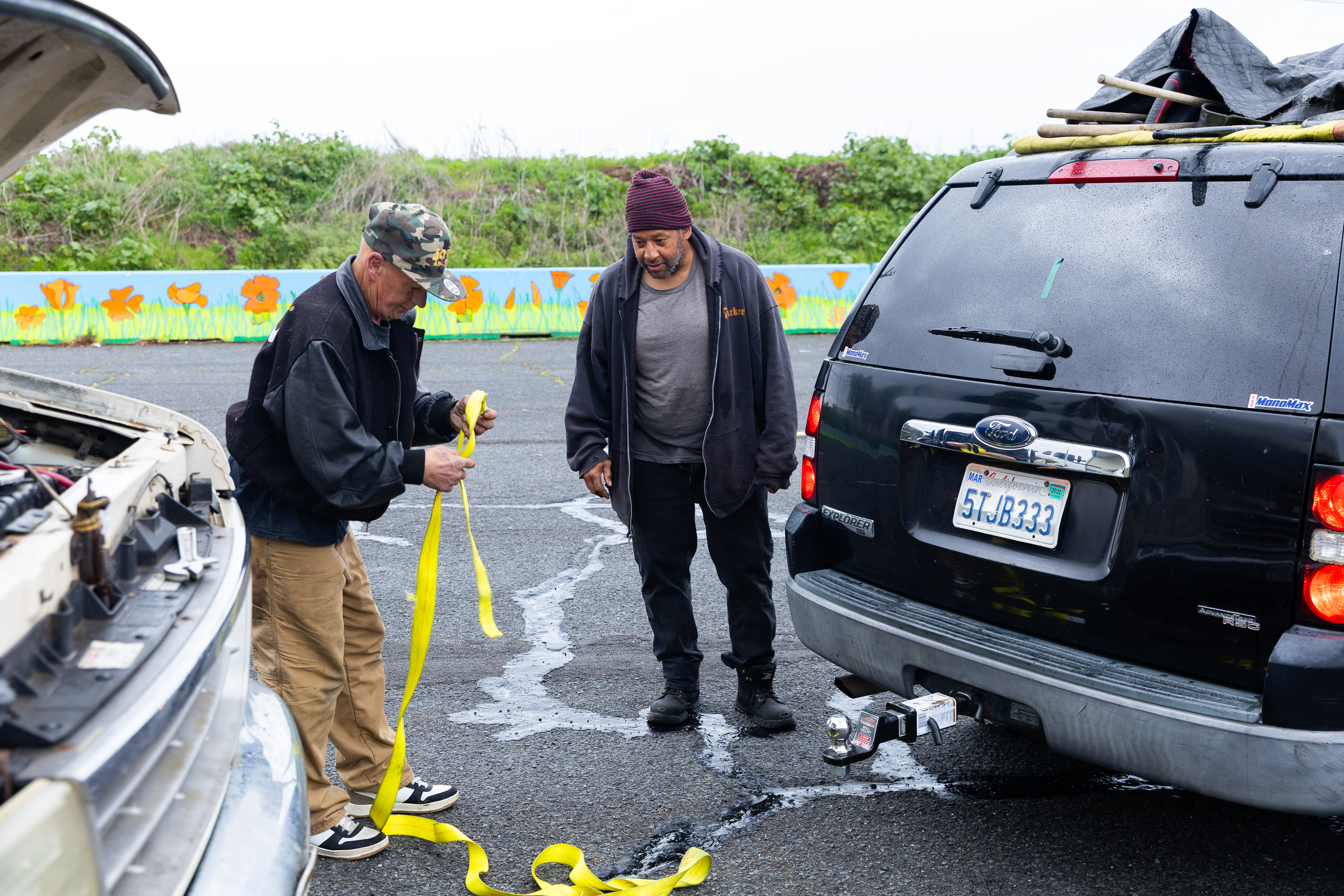 Two men are standing near a black SUV and a car with an open hood. One man is holding a yellow strap. There's a painted wall and greenery behind them.