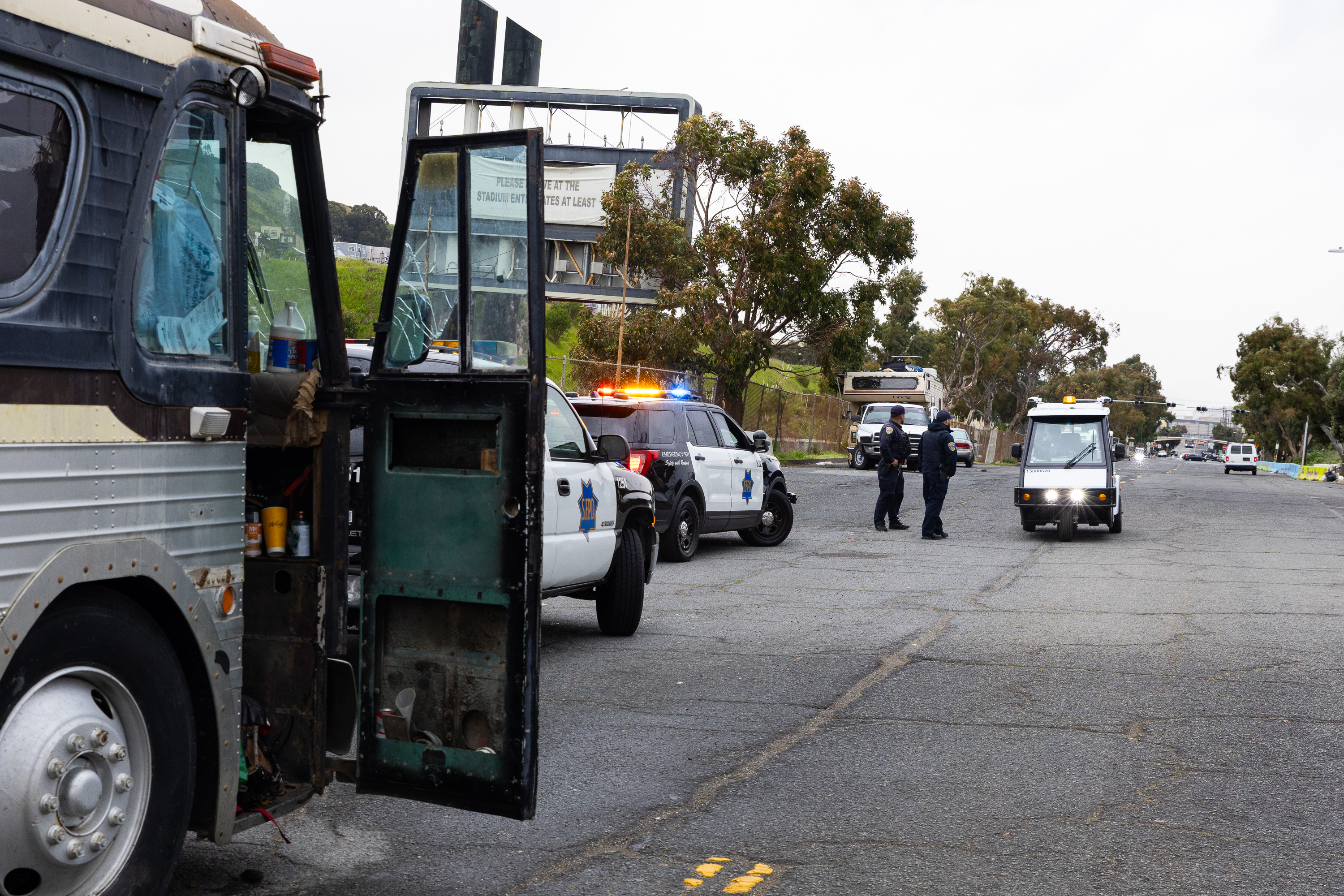 The image shows a street scene with an old bus with an open door on the left. Police cars with lights on and officers are nearby. Other vehicles are in the distance.