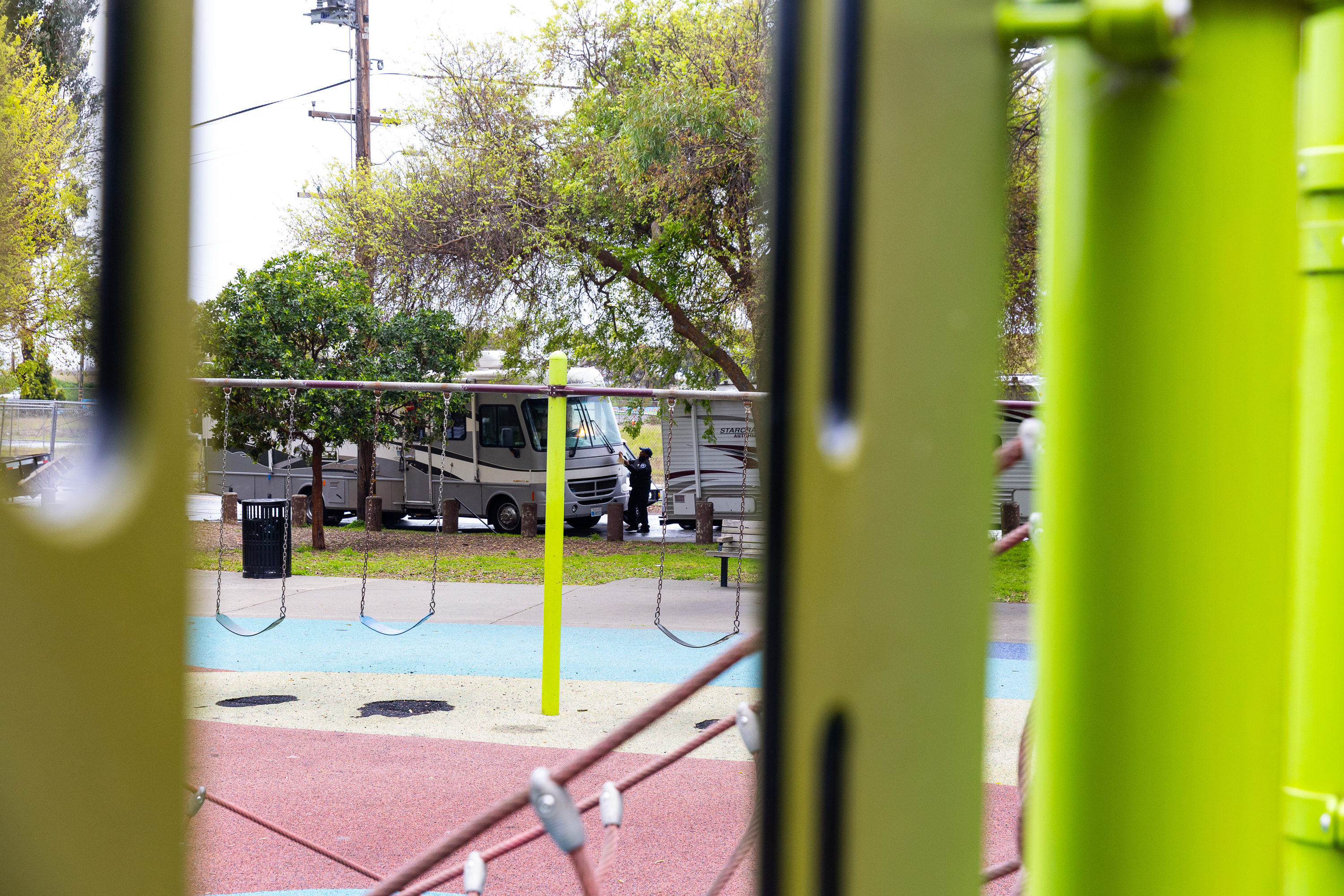 The image shows a playground with empty swings. In the background, there's a person near two RVs, surrounded by trees and more playground equipment.