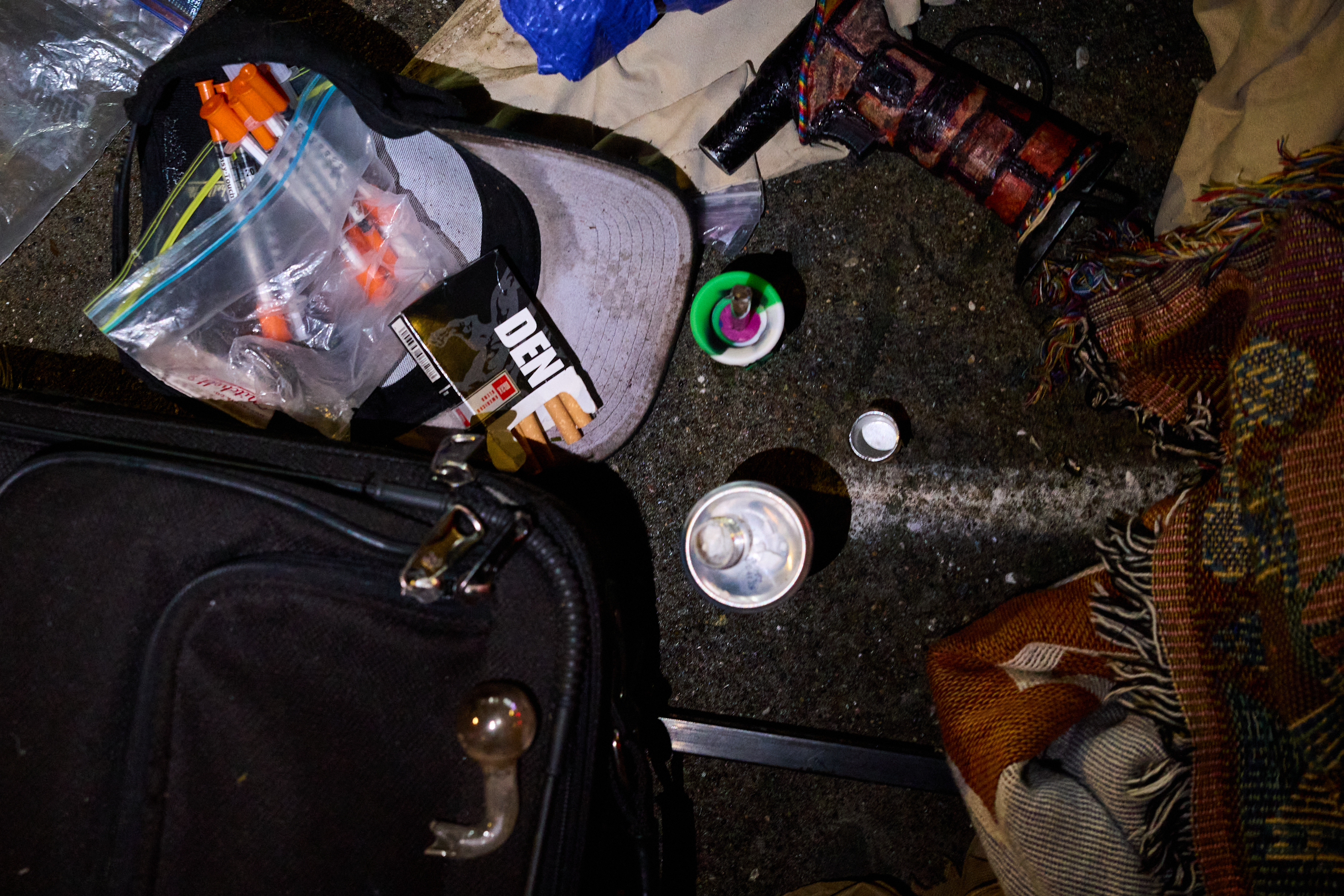 The image shows a cluttered ground with a black suitcase, a cap filled with syringes, a can, a colorful woven item, and other miscellaneous objects.