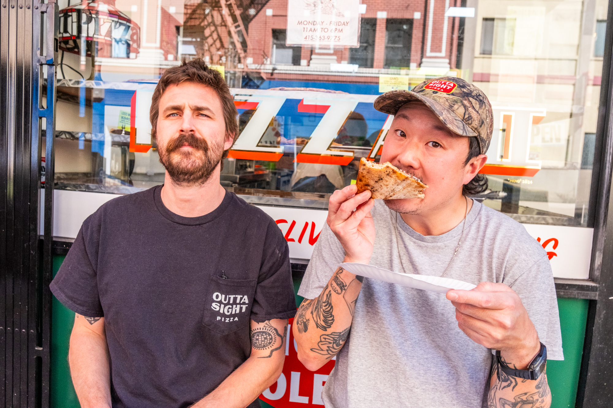 Two men sit outside a pizzeria. The left man looks at the camera, while the right man, wearing a cap, bites into a slice of pizza on a paper plate.