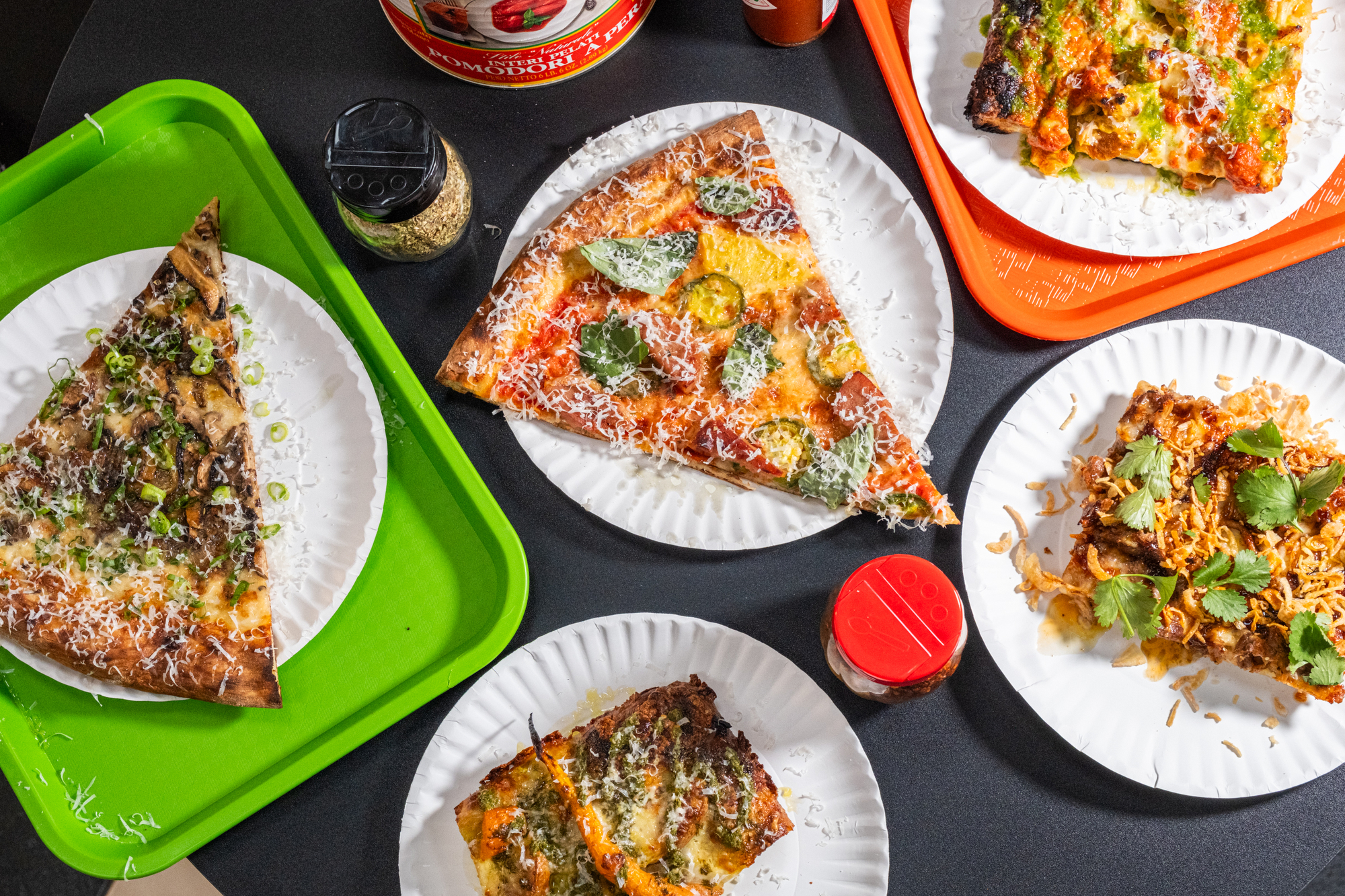 The image shows various slices of pizza on paper plates, each with different toppings like cheese, herbs, and vegetables. They are placed on colorful trays, with condiments nearby.