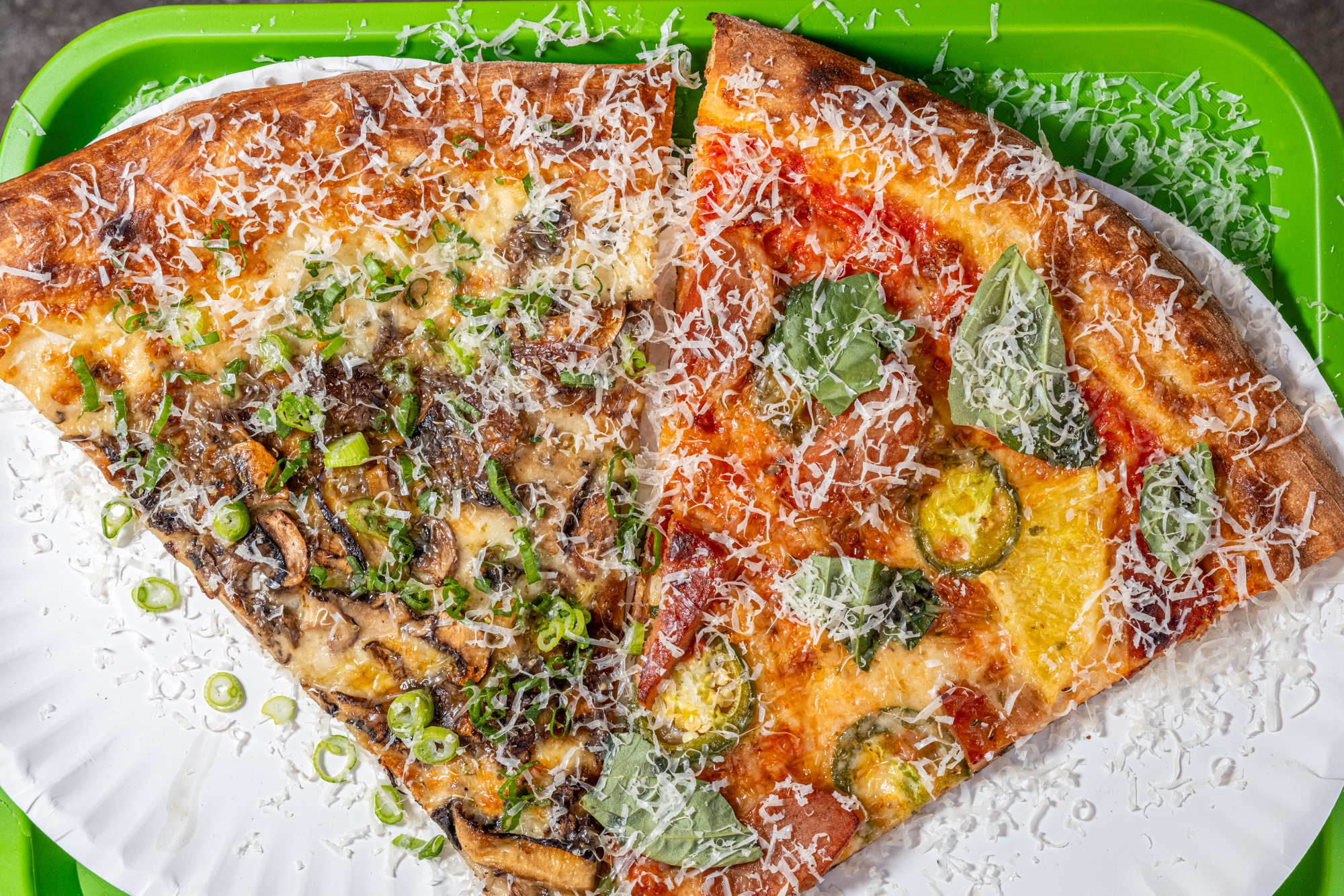 Two pizza slices on a white paper plate, one topped with mushrooms and green onions, the other with basil and jalapeños, both sprinkled with cheese.