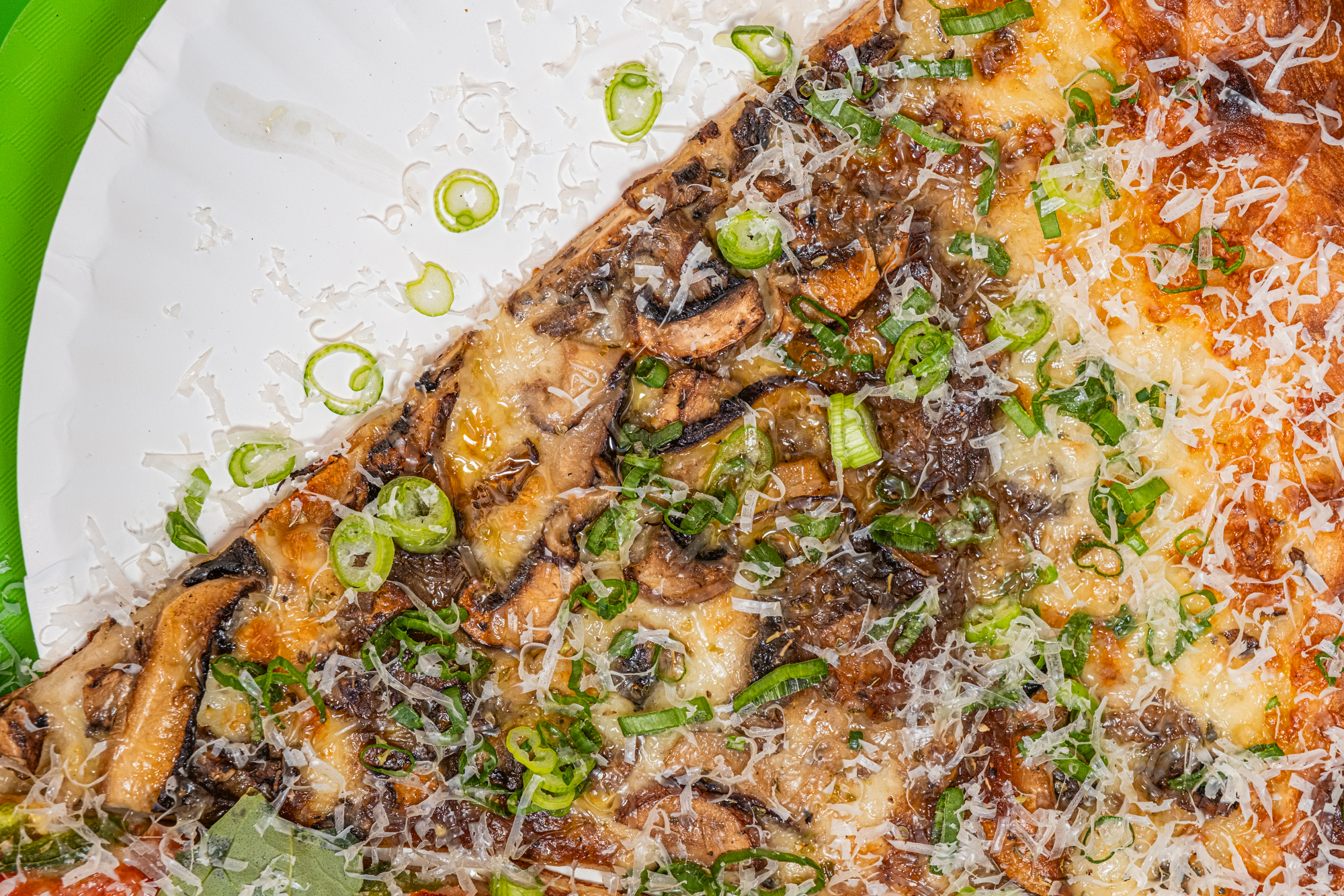 A pizza slice on a plate is topped with mushrooms, green onions, and grated cheese. The plate is white with a green rim.