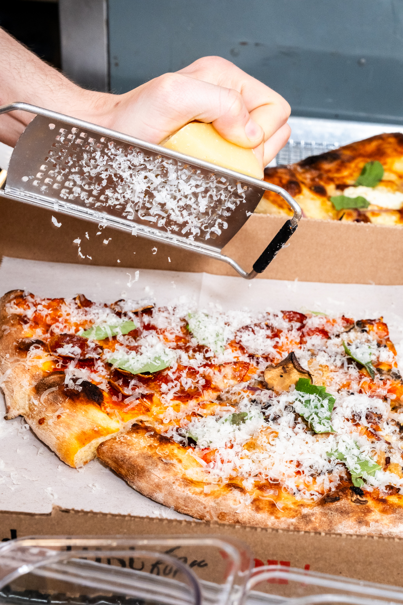 A hand grates cheese onto a pizza topped with pepperoni and basil inside a cardboard box. The pizza has a golden-brown crust.