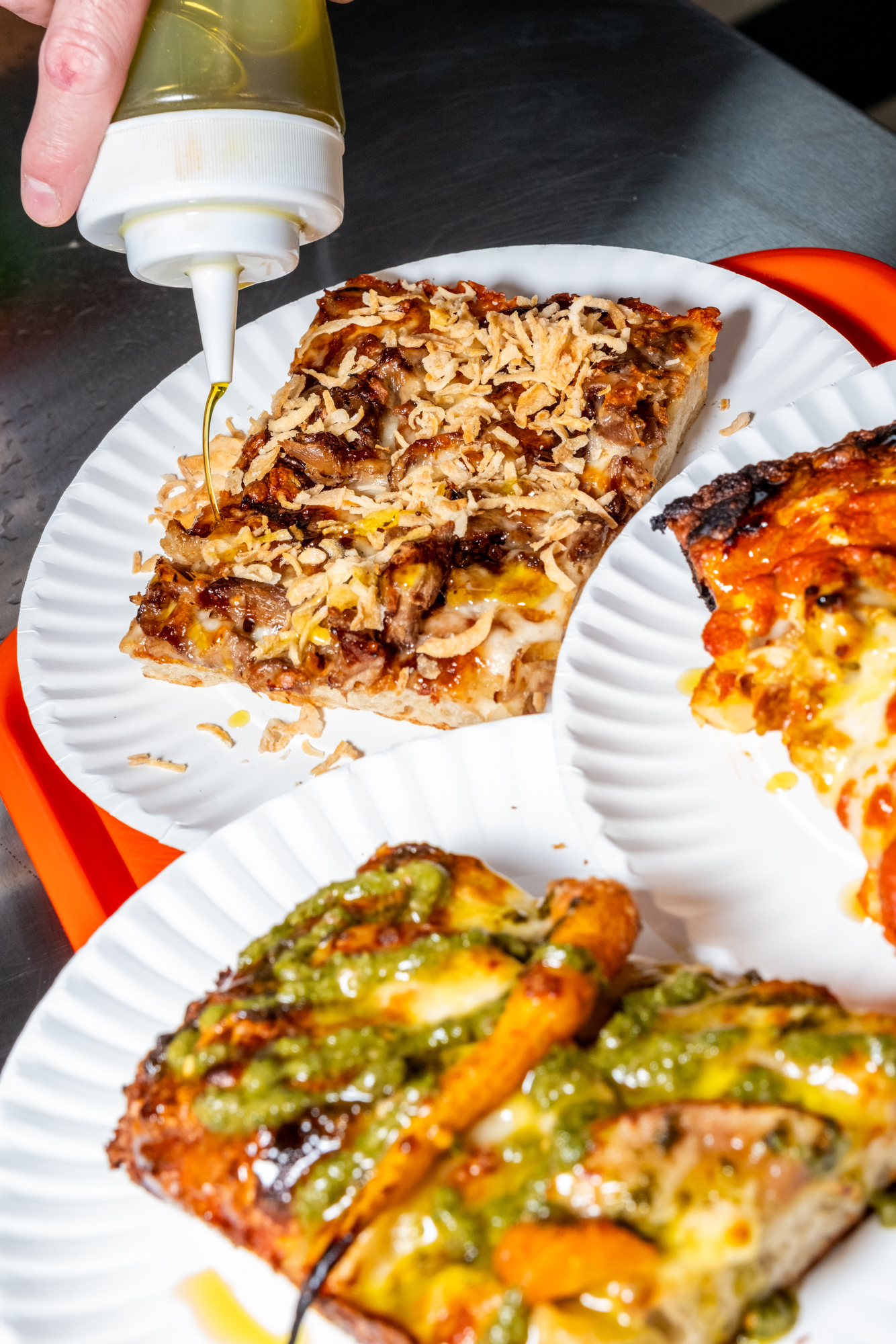 A hand drizzles oil on a pizza slice with crispy onions. Two other slices, one with pesto, rest on white paper plates over a red tray.