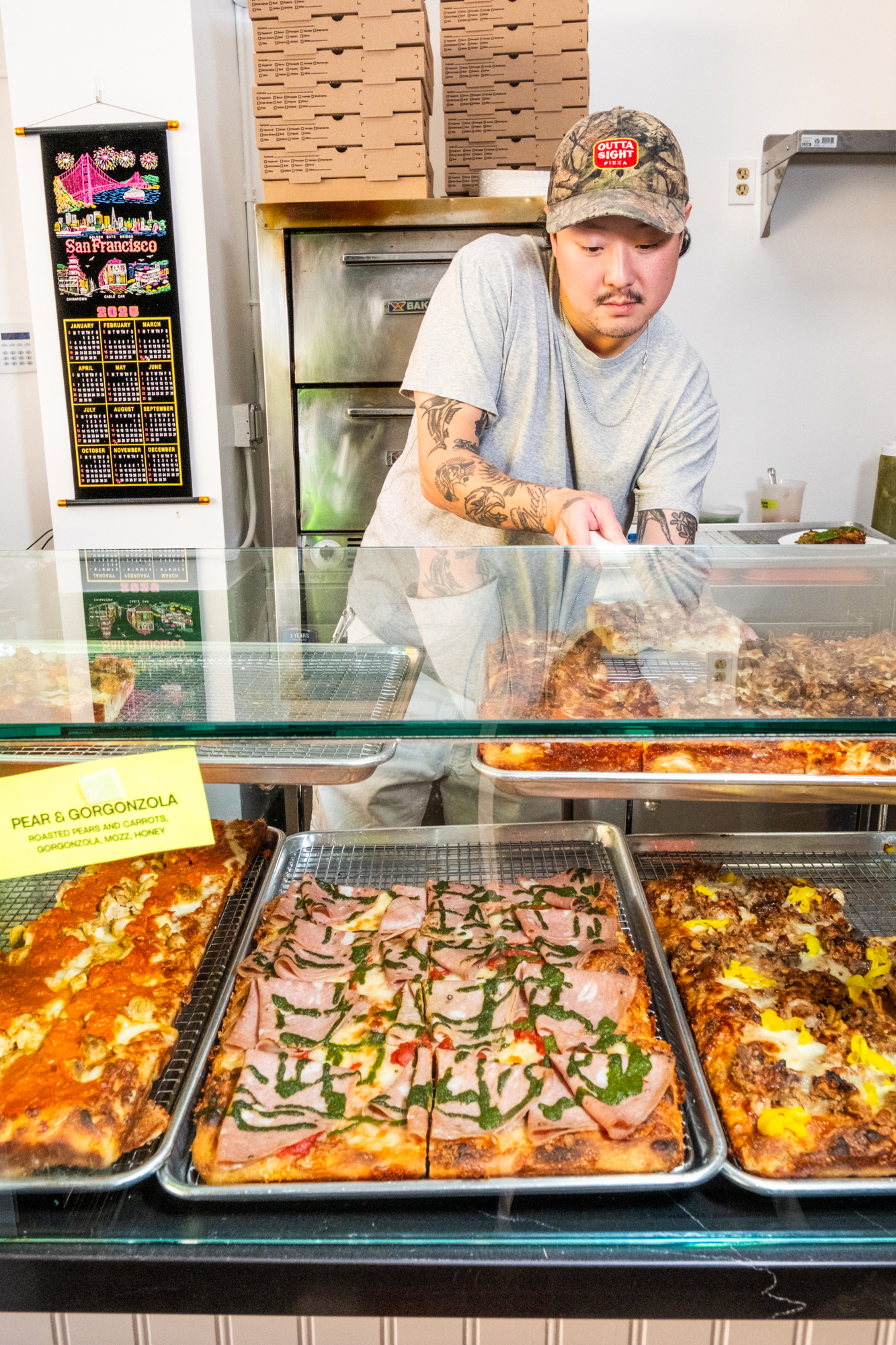 A person in a cap serves pizza from a display with varieties like pear and gorgonzola. Pizza boxes and a San Francisco calendar hang nearby.