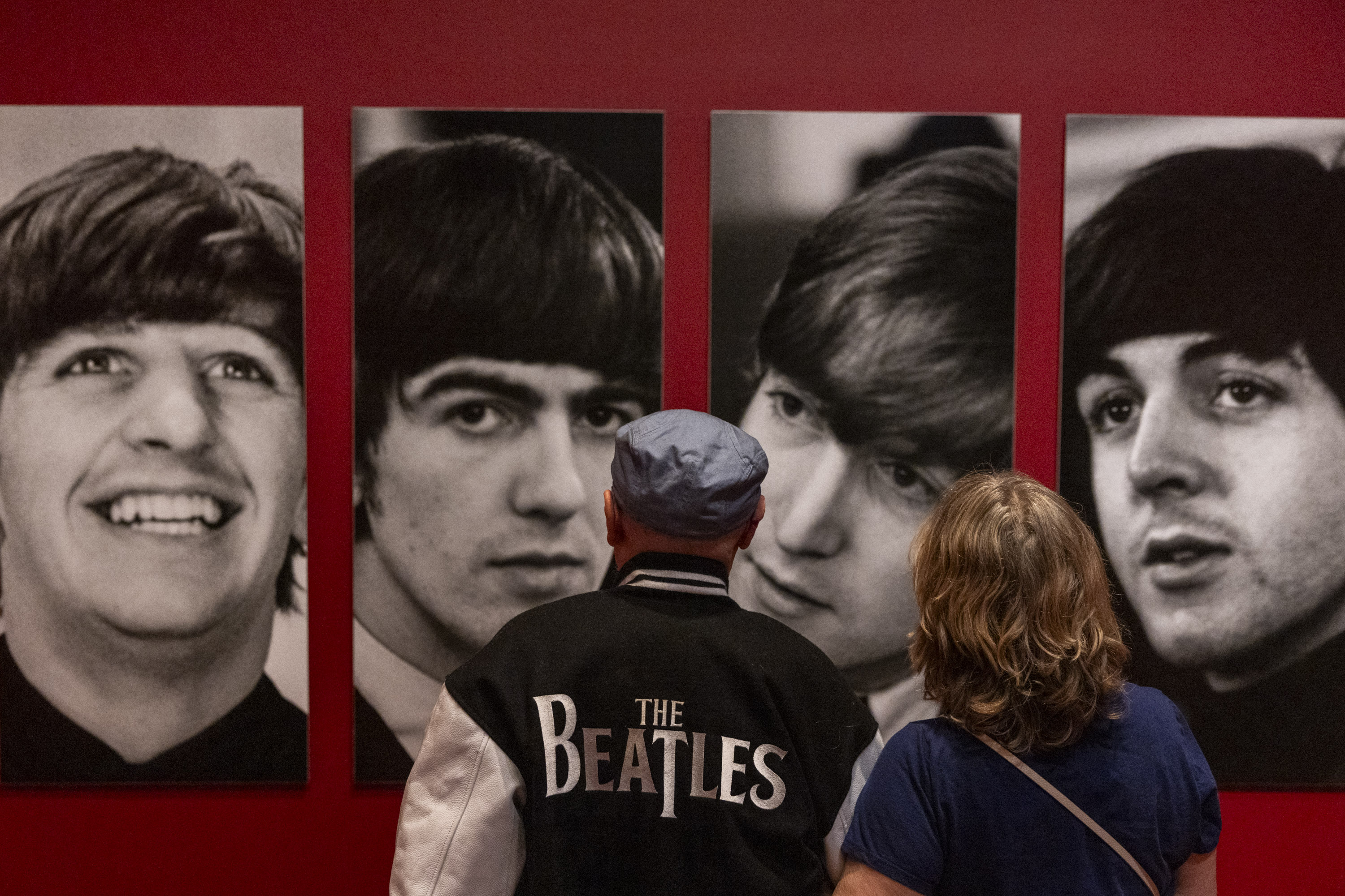 Two people stand in front of black and white portraits of four individuals displayed on a red wall. The back of one person's jacket reads "The Beatles."