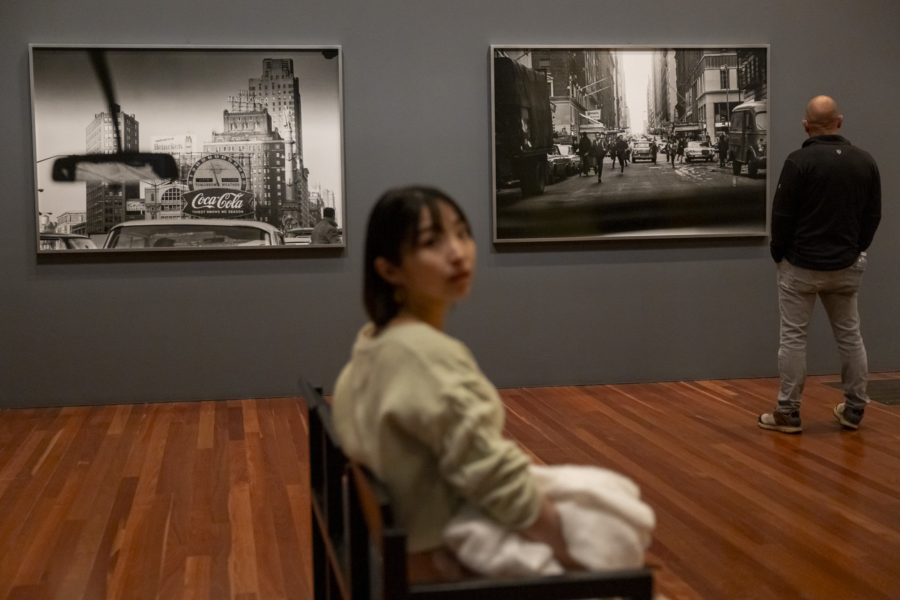 The image shows two people in an art gallery viewing black-and-white cityscape photos. One person is seated and another, standing, faces the artwork.