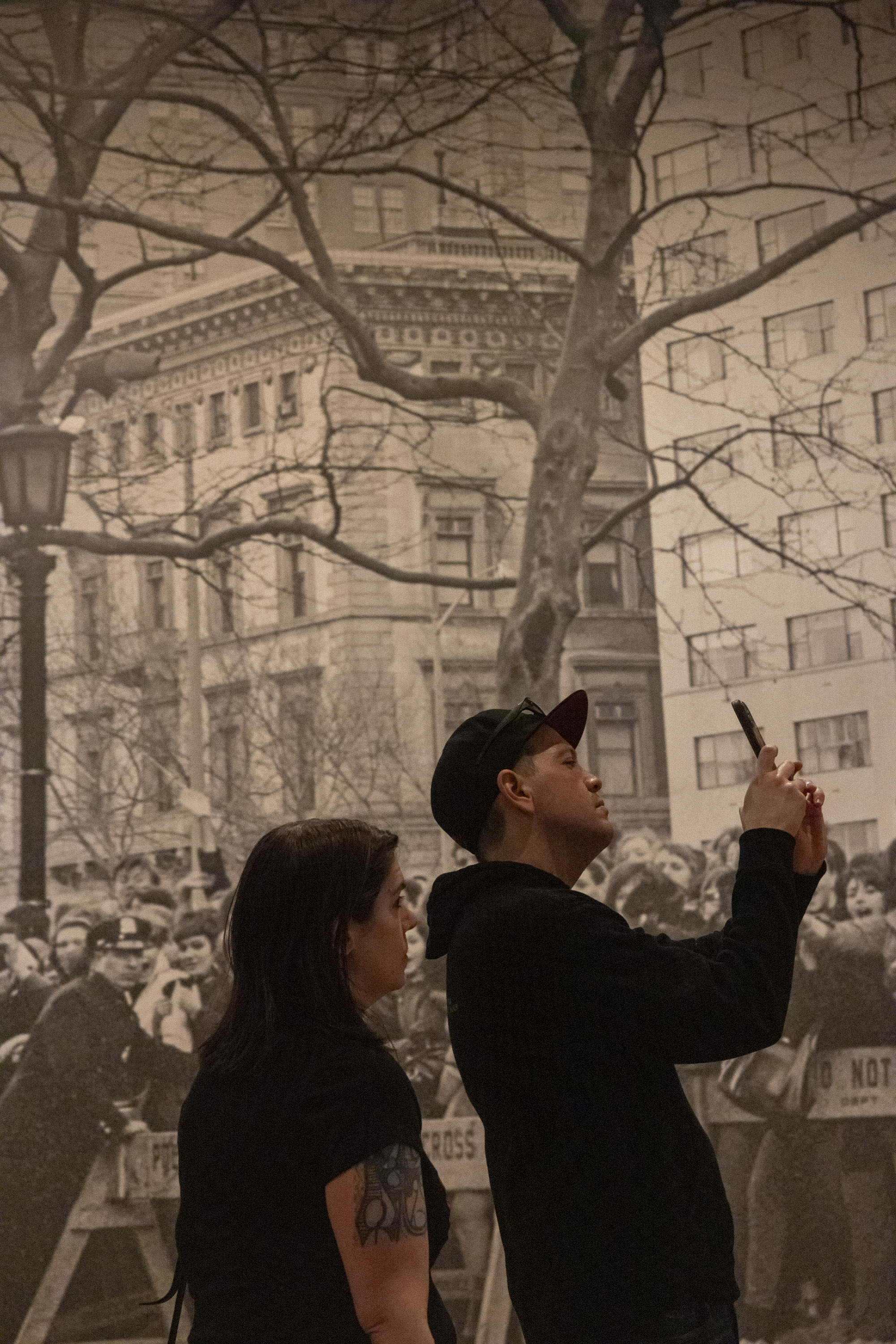 A man in a cap holds up a phone, taking a picture in front of a large black-and-white mural depicting a crowd, buildings, and bare trees. A woman stands beside him.