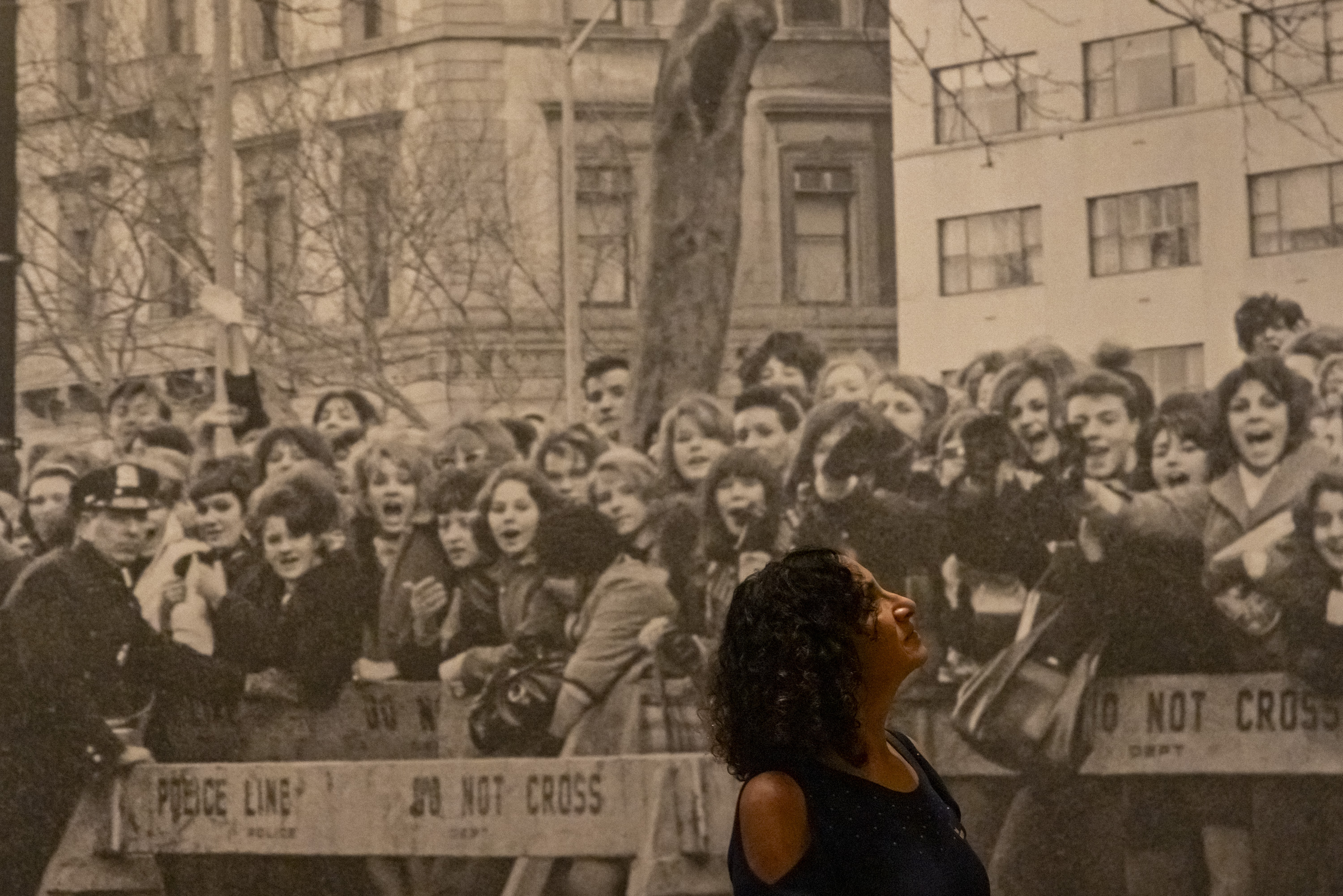 A woman in a dark top stands in front of a large black-and-white photo of a historical crowd scene, including a police officer and excited onlookers.