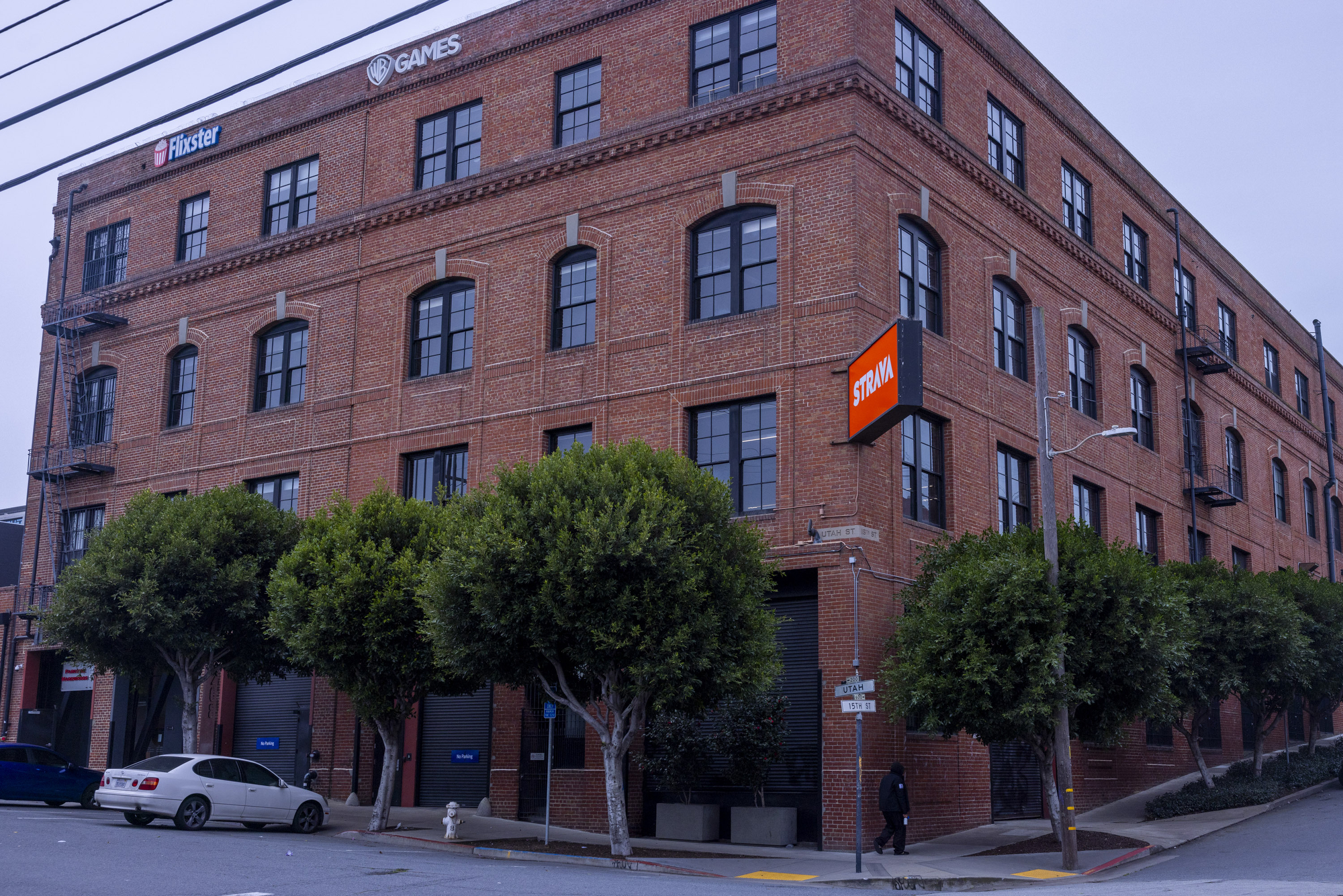 A large brick building houses several companies, including Strava and WB Games, as indicated by signs. Trees line the street along parked cars.