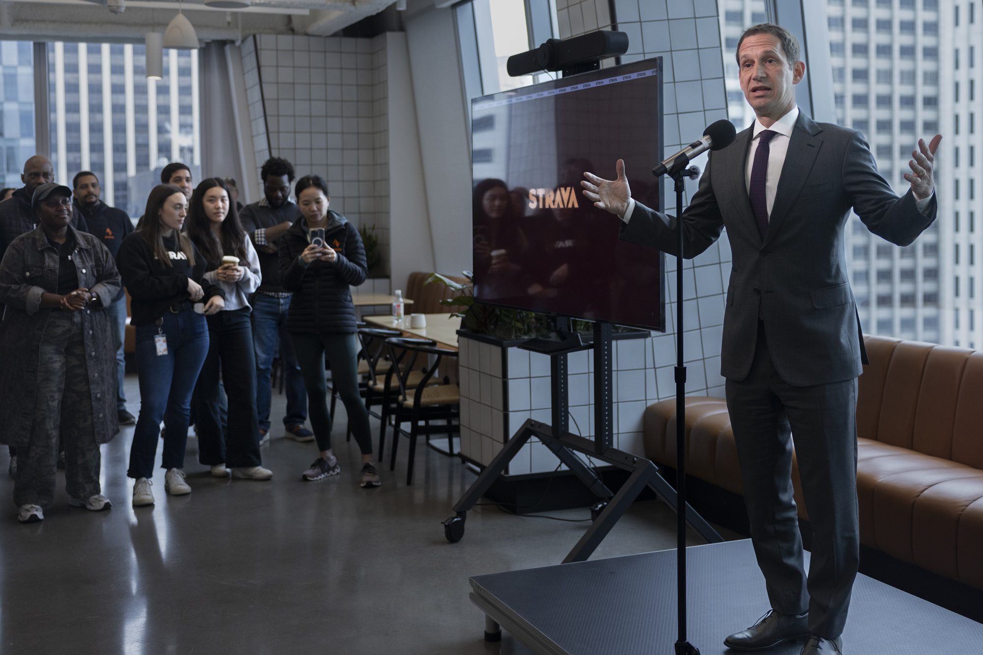 A man in a suit speaks into a microphone on a small stage, with an audience of diverse people standing and observing. A large screen is next to him displaying &quot;Strava.&quot;