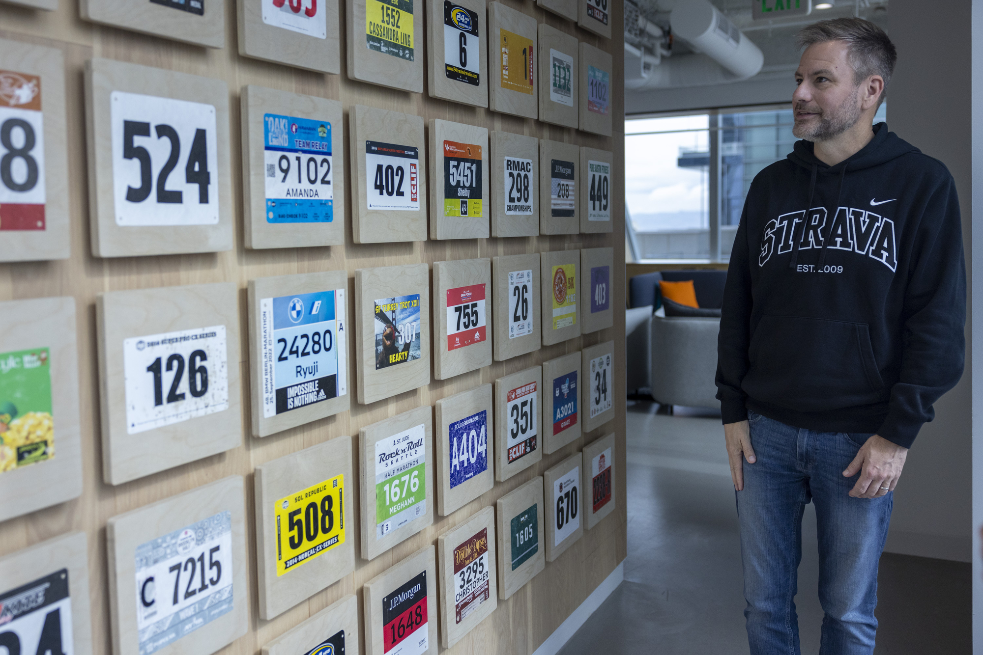 A man in a black hoodie with "STRAVA" on it stands in an office, looking at a wall covered in framed race bibs displayed in a neat grid.