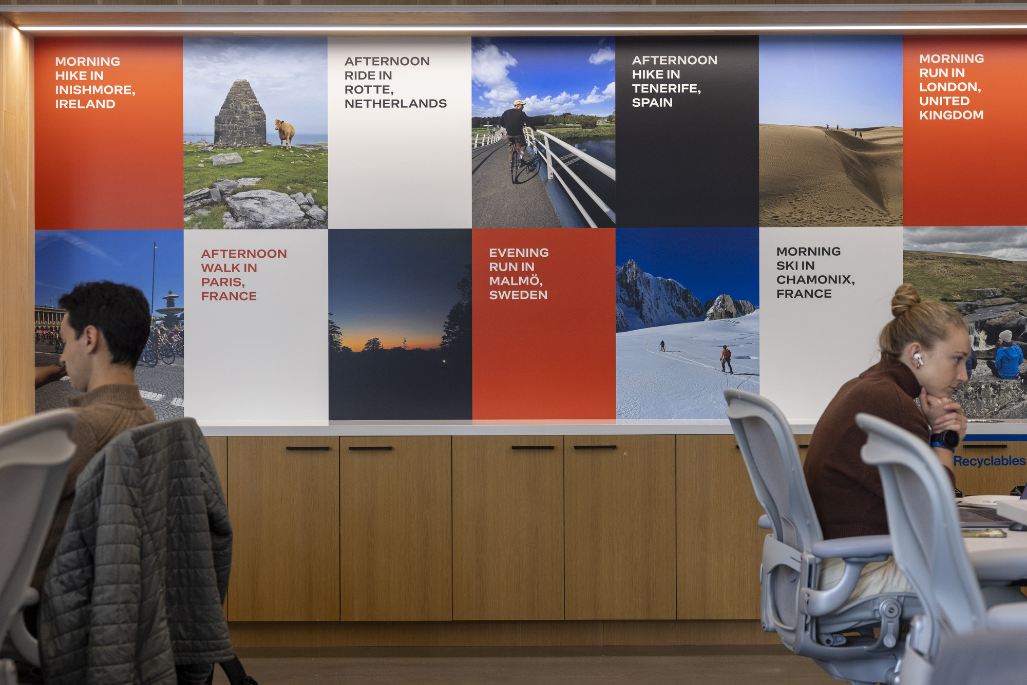 A wall displays images and text of outdoor activities worldwide, like hiking and skiing. Below are wooden cabinets. Two people work in office chairs.