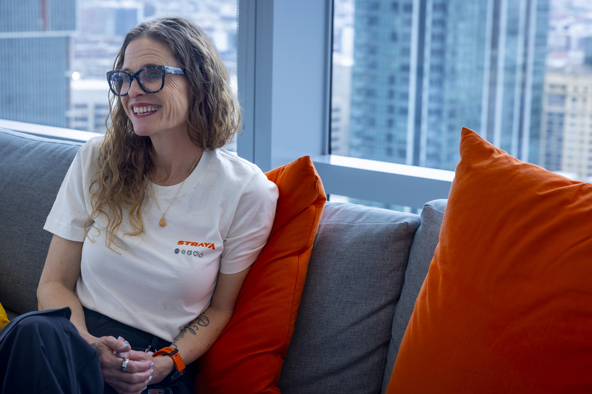 A smiling woman with curly hair and glasses sits on a couch with orange pillows, wearing a white shirt with &quot;STRAVA&quot; on it. A cityscape is visible through the window.