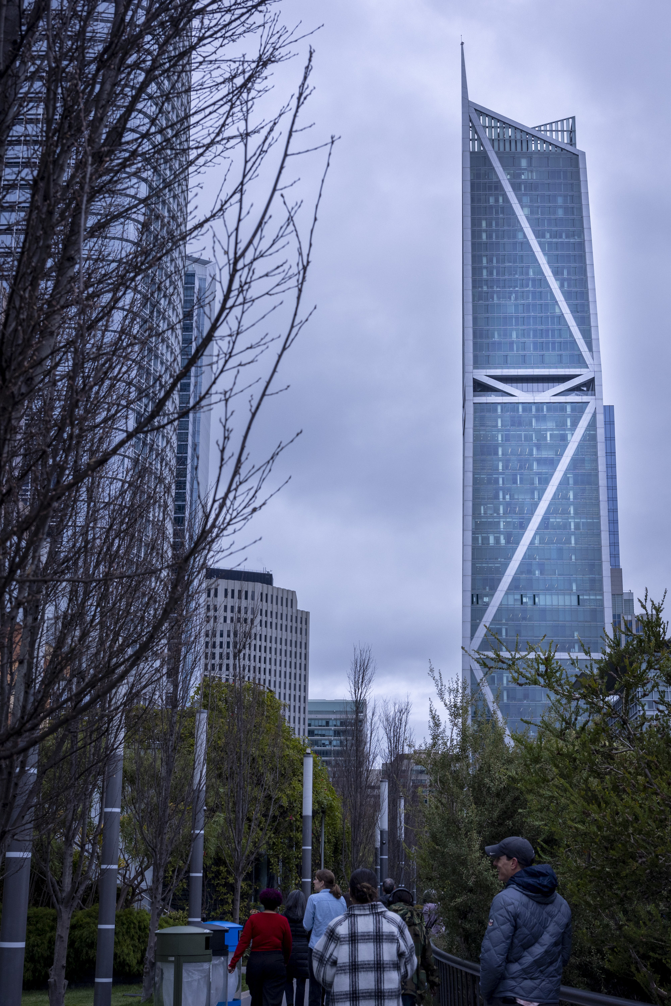 The image shows a group of people walking along a tree-lined path with a modern, angular skyscraper in the background under a cloudy sky.