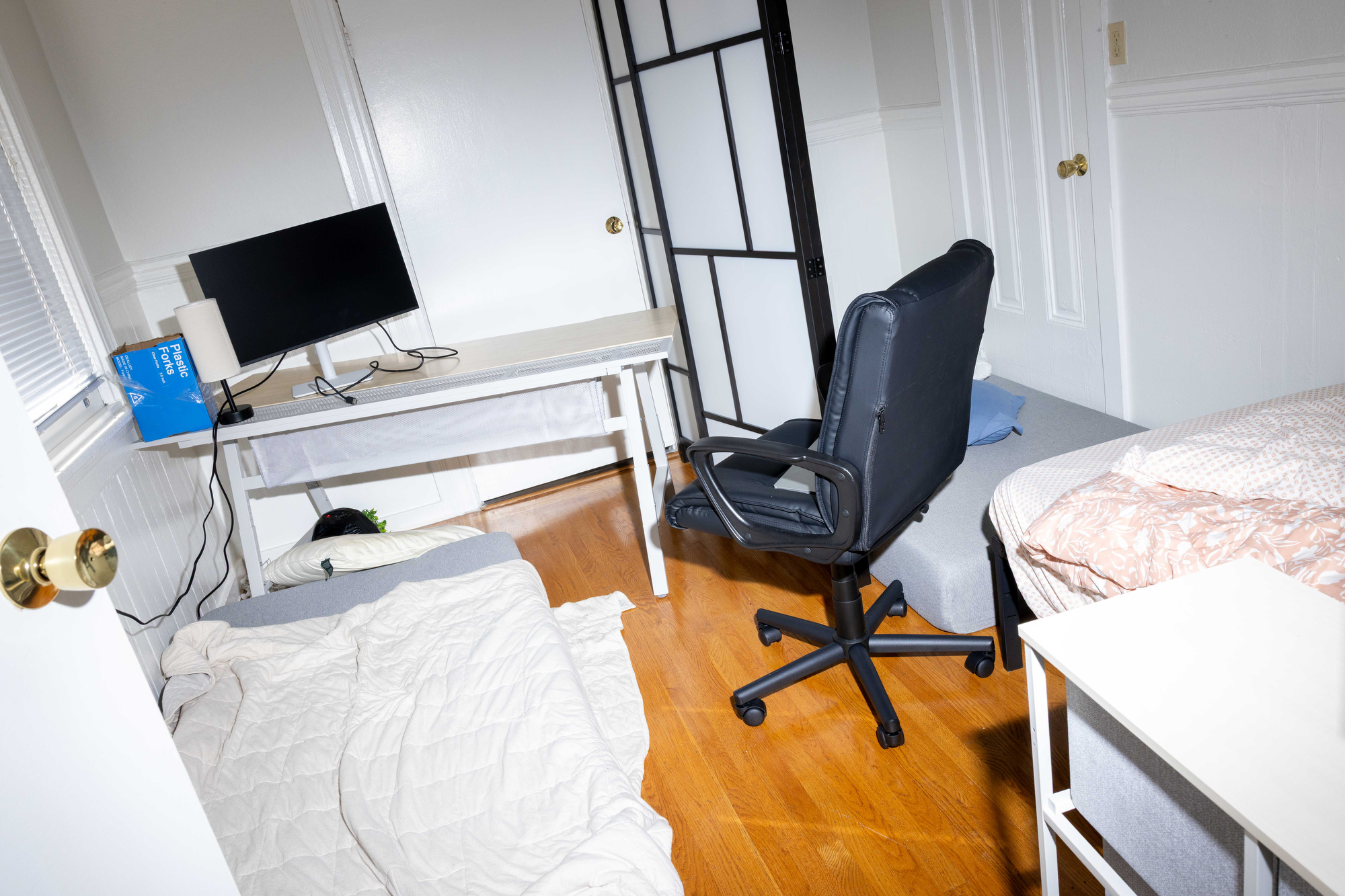 The room has a desk with a computer monitor, a rolling chair, two beds with white and patterned bedding, and a hardwood floor.