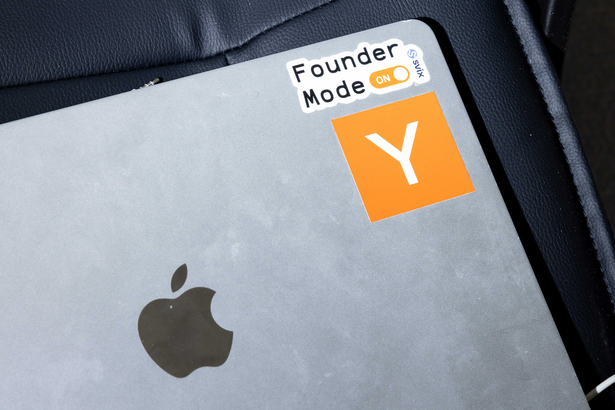 A silver laptop is on a dark surface, featuring an Apple logo and stickers saying &quot;Founder Mode ON&quot; and an orange square with a white &quot;Y.&quot;