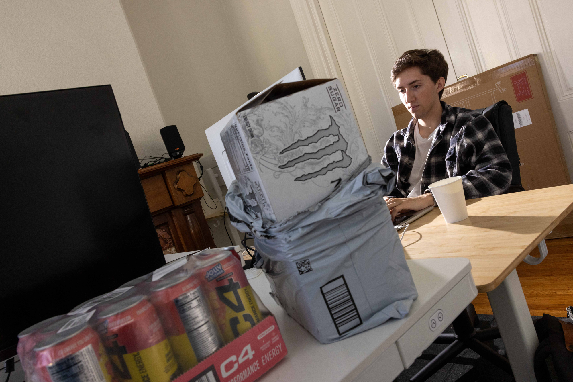 A person sits at a desk using a computer, with a soft drink can pack and a paper cup nearby. A TV and packages are in the background.