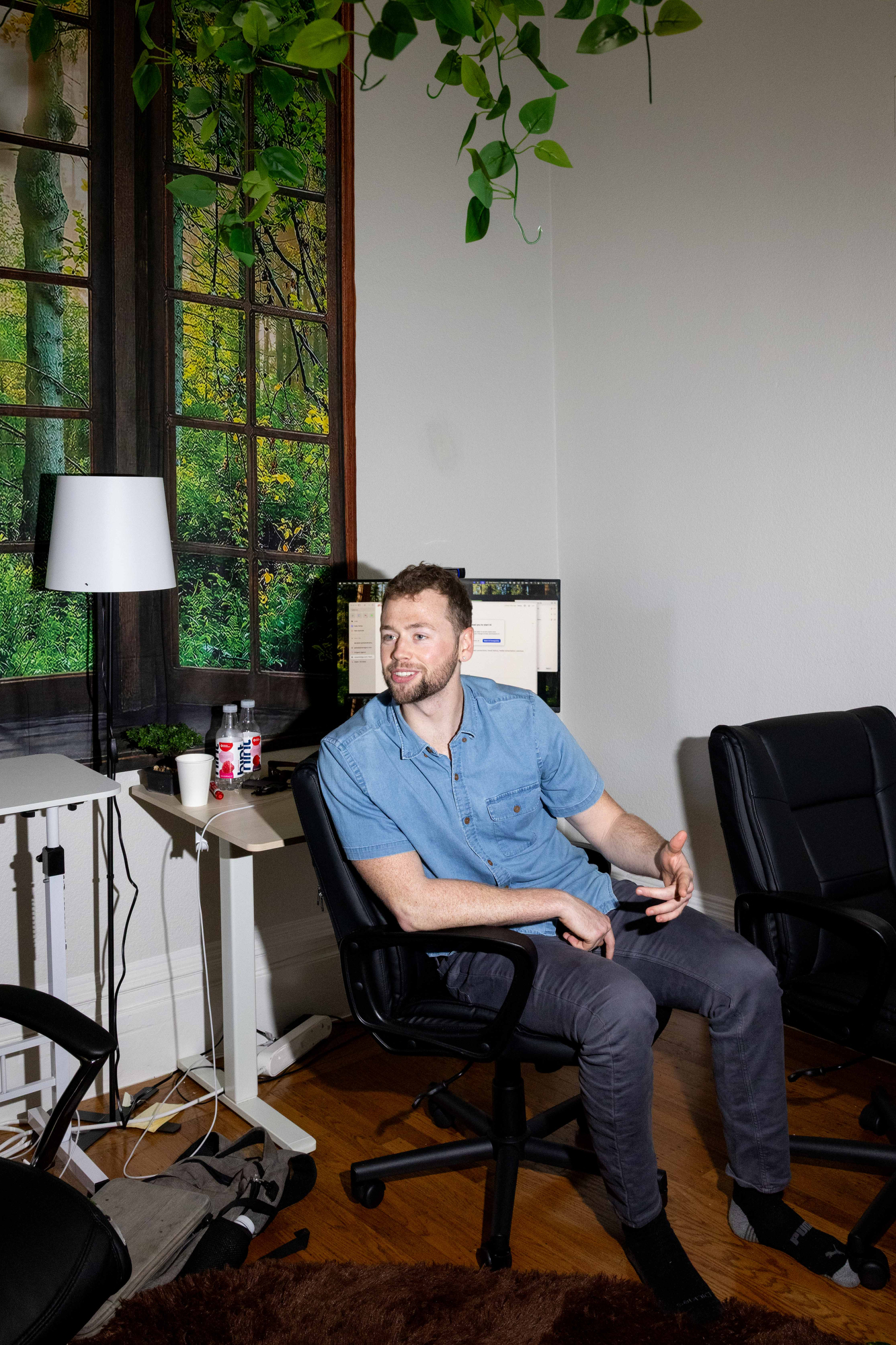 A man in a blue shirt sits on a chair in a home office with a desk, lamp, and computer. A leafy plant hangs overhead, and a window with a forest view is behind.