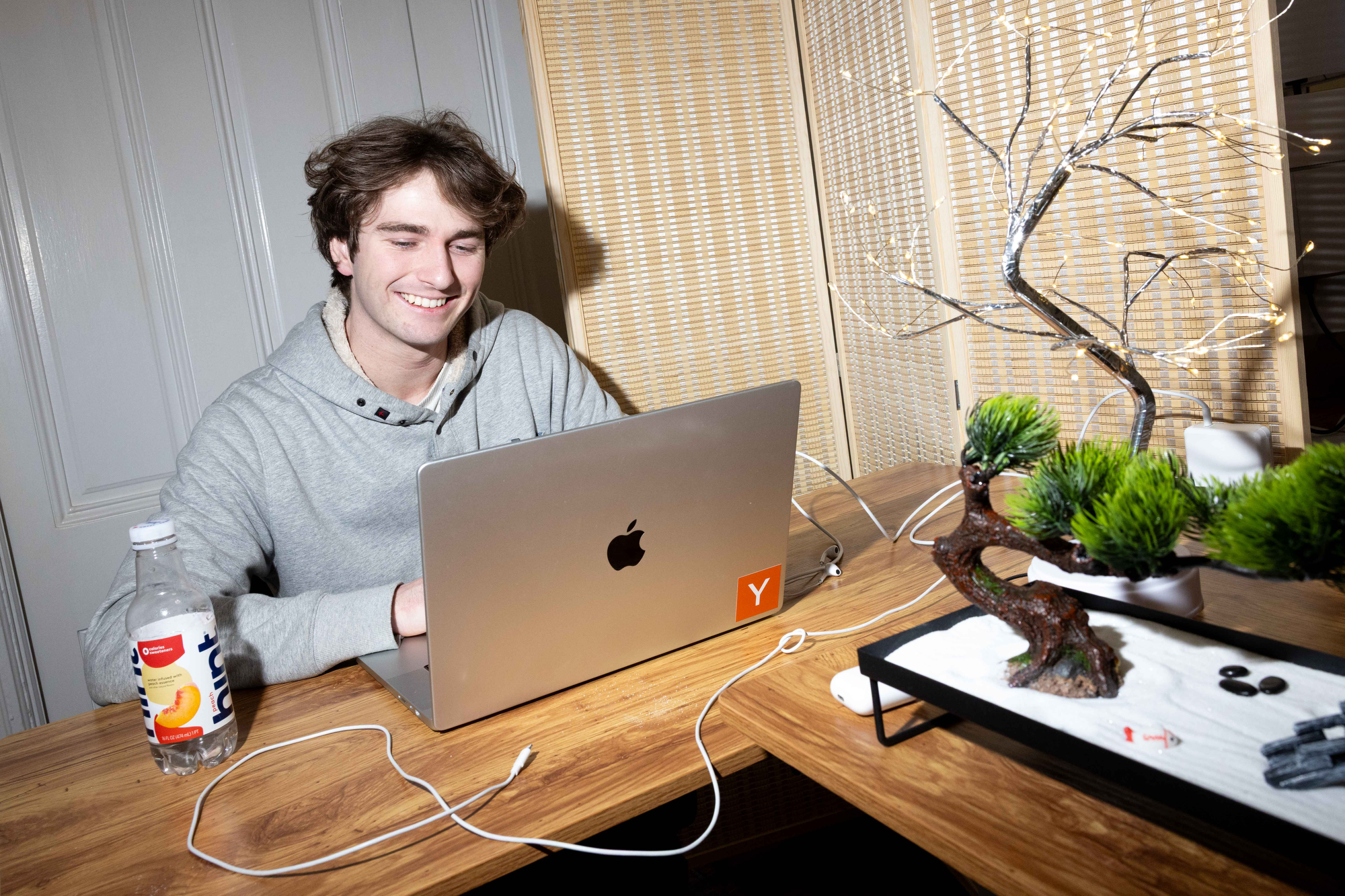 A person in a gray hoodie smiles while using a laptop on a wooden table. Nearby, there’s a bottle of water and a decorative bonsai tree with lights.