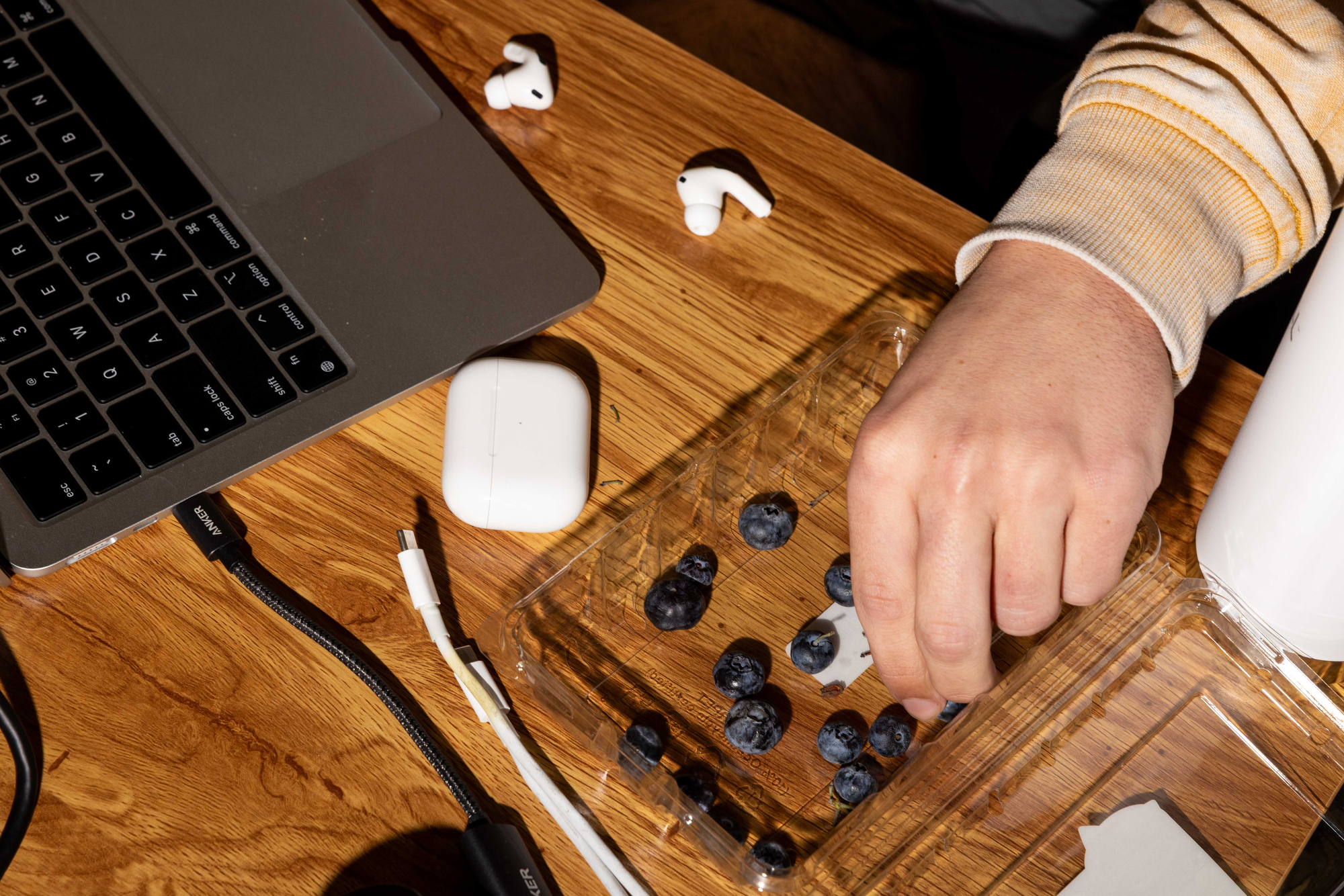 A hand picks blueberries from a plastic container on a wooden table. Nearby are earbuds, a laptop, and charging cables.