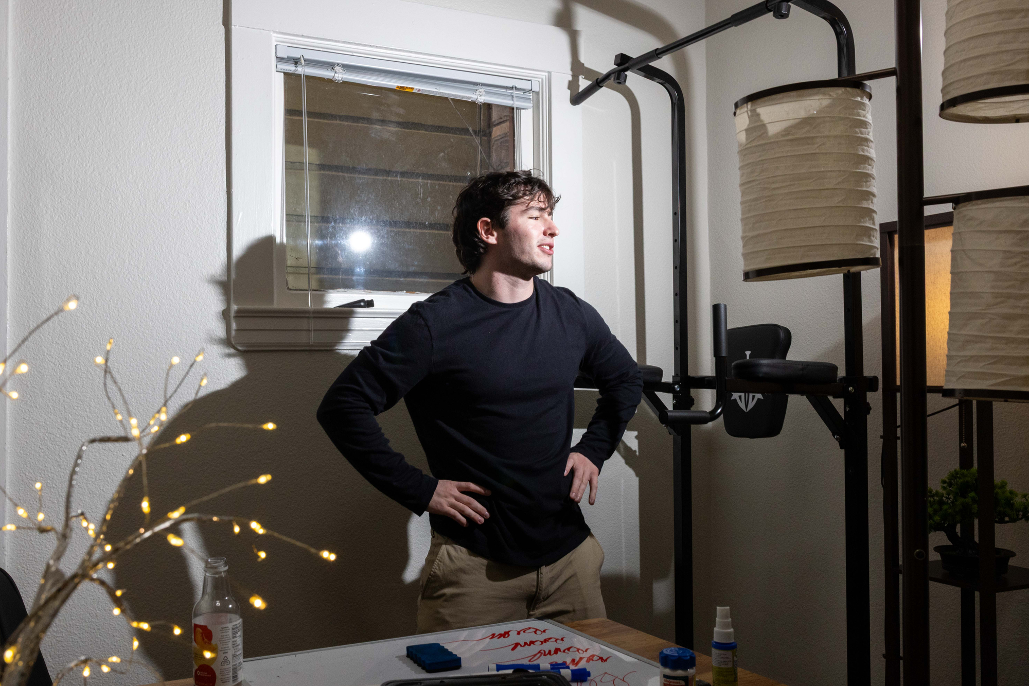 A person in a black shirt stands with hands on hips near a window. A table with markers and paper is in the foreground, and fairy lights and lamps add ambiance.