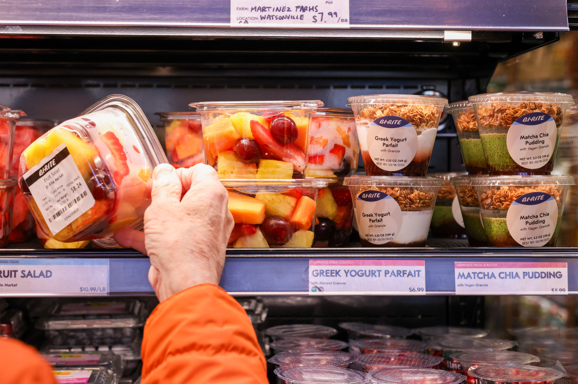 A person in an orange sleeve reaches for a fruit salad container on a shelf, next to Greek yogurt parfaits and matcha chia puddings, all neatly displayed.