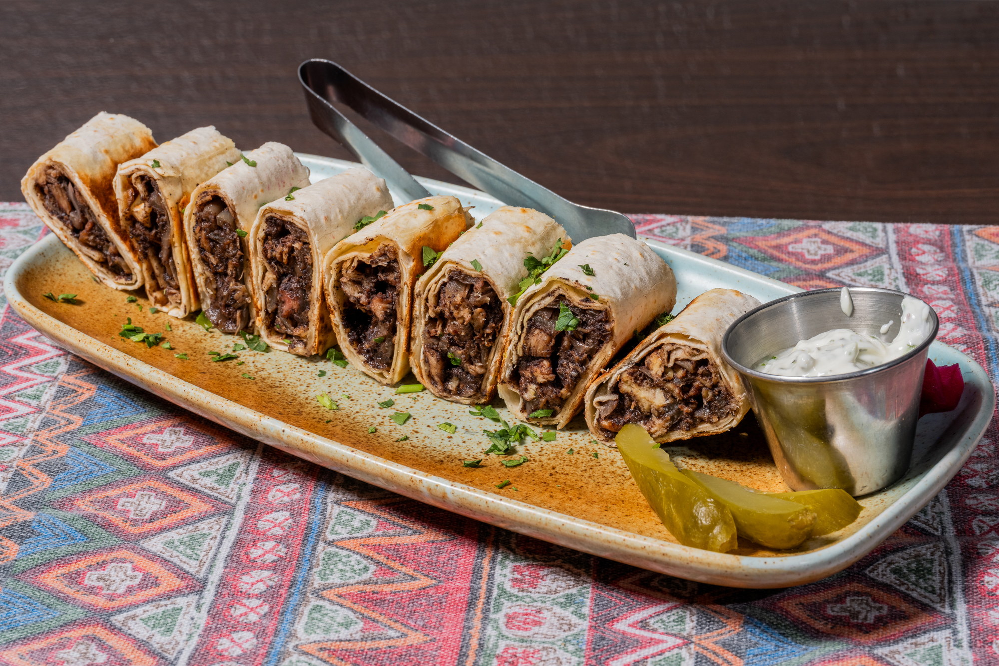 The image shows sliced wraps filled with meat, arranged on a plate with parsley garnish. A metal cup holds a creamy sauce, next to pickled vegetables.