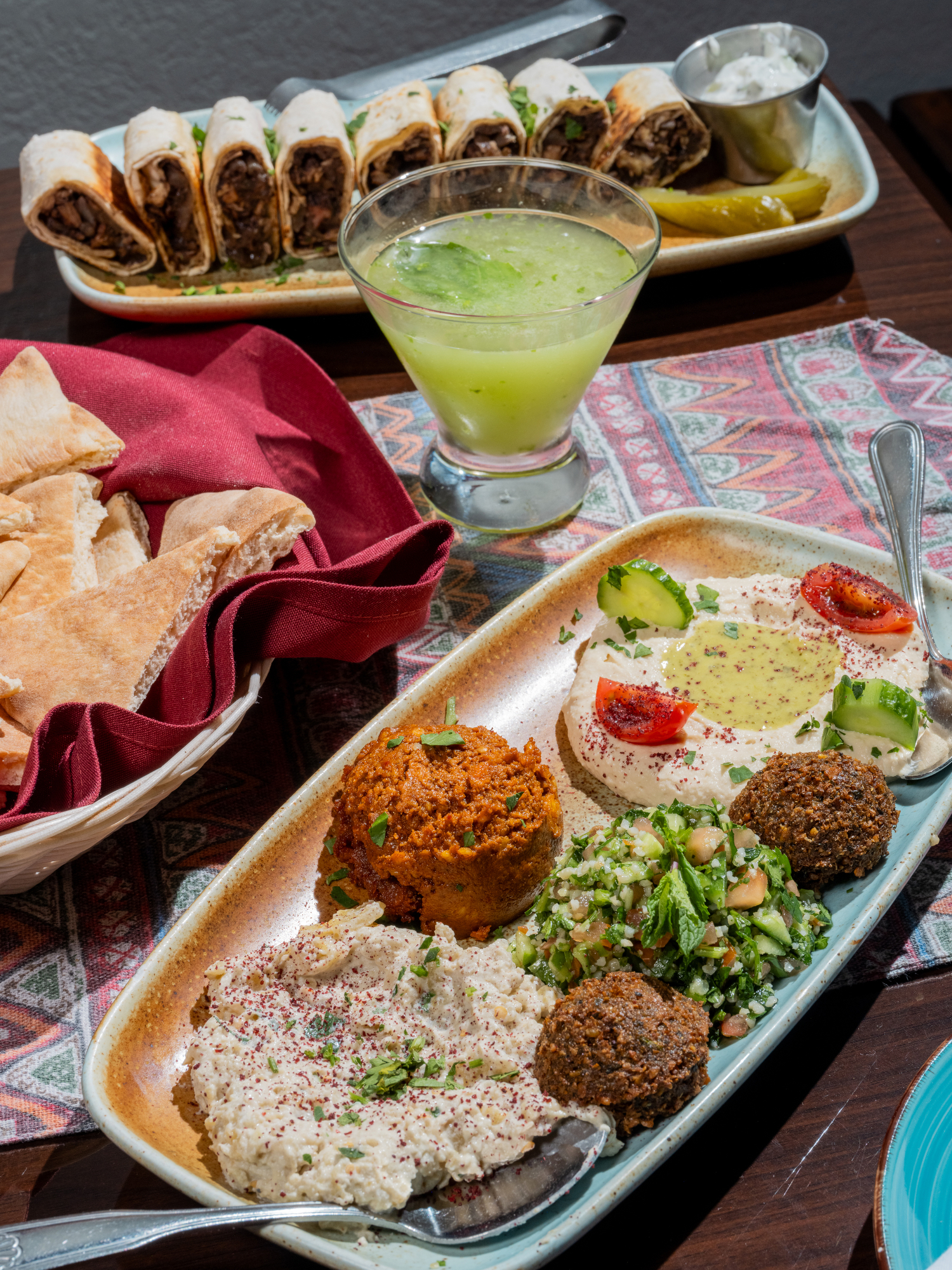 A platter with hummus, falafel, baba ghanoush, and tabbouleh, alongside pita bread. In the background, there are rolled appetizers and a green drink.