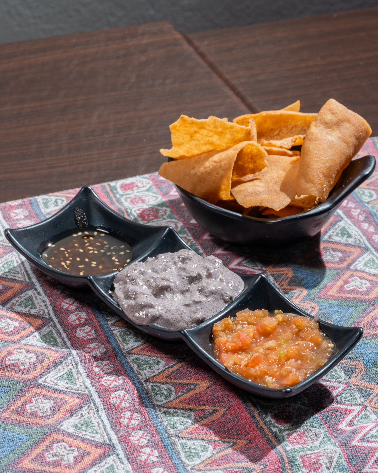 The image shows a bowl of crispy chips beside a tray with three sauces: dark soy sauce with seeds, gray thick paste, and chunky tomato salsa, on a patterned cloth.