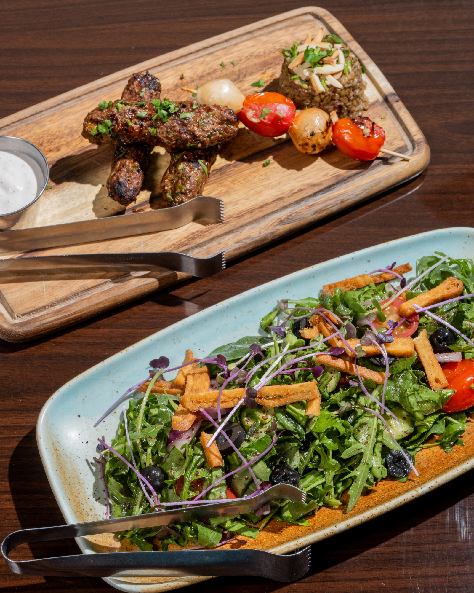 The image shows a wooden board with grilled meat skewers, vegetables, and rice, and a plate of leafy salad with olives, tomatoes, and crispy strips.