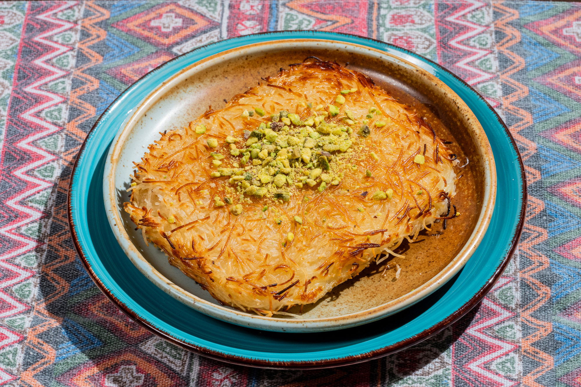 A baked dessert topped with crushed pistachios is on a blue-edged plate. The background is a colorful, geometric-patterned fabric.