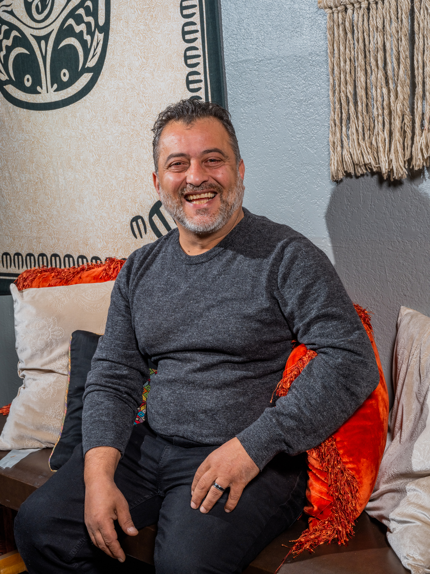 A smiling person is sitting on a bench with colorful pillows, wearing a gray sweater. The background features a patterned tapestry and macrame art.