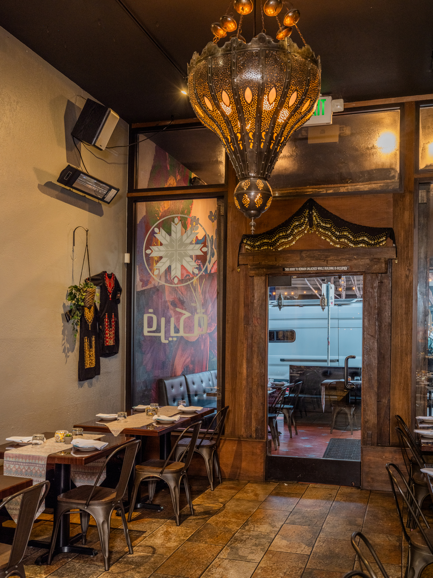 The image shows a cozy restaurant with wooden tables and metal chairs. A large, ornate chandelier hangs above. The walls have decorative art and traditional garments.
