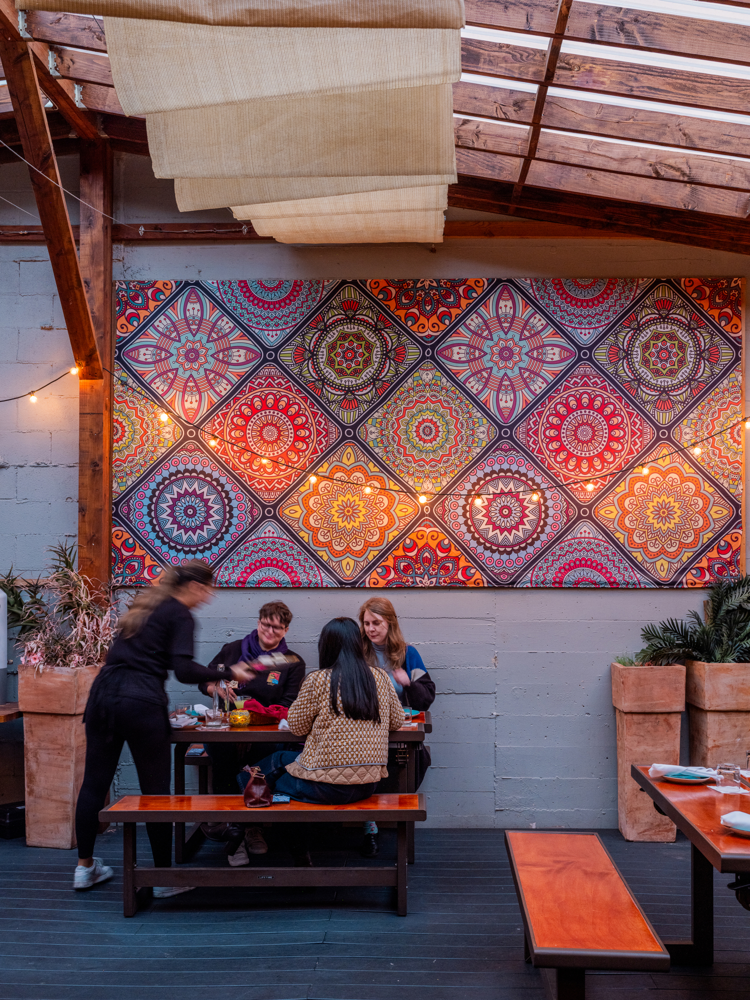 A group of people dine at a table under a wooden canopy, with colorful, intricate patterns on the wall behind them. A server is attending to the diners.